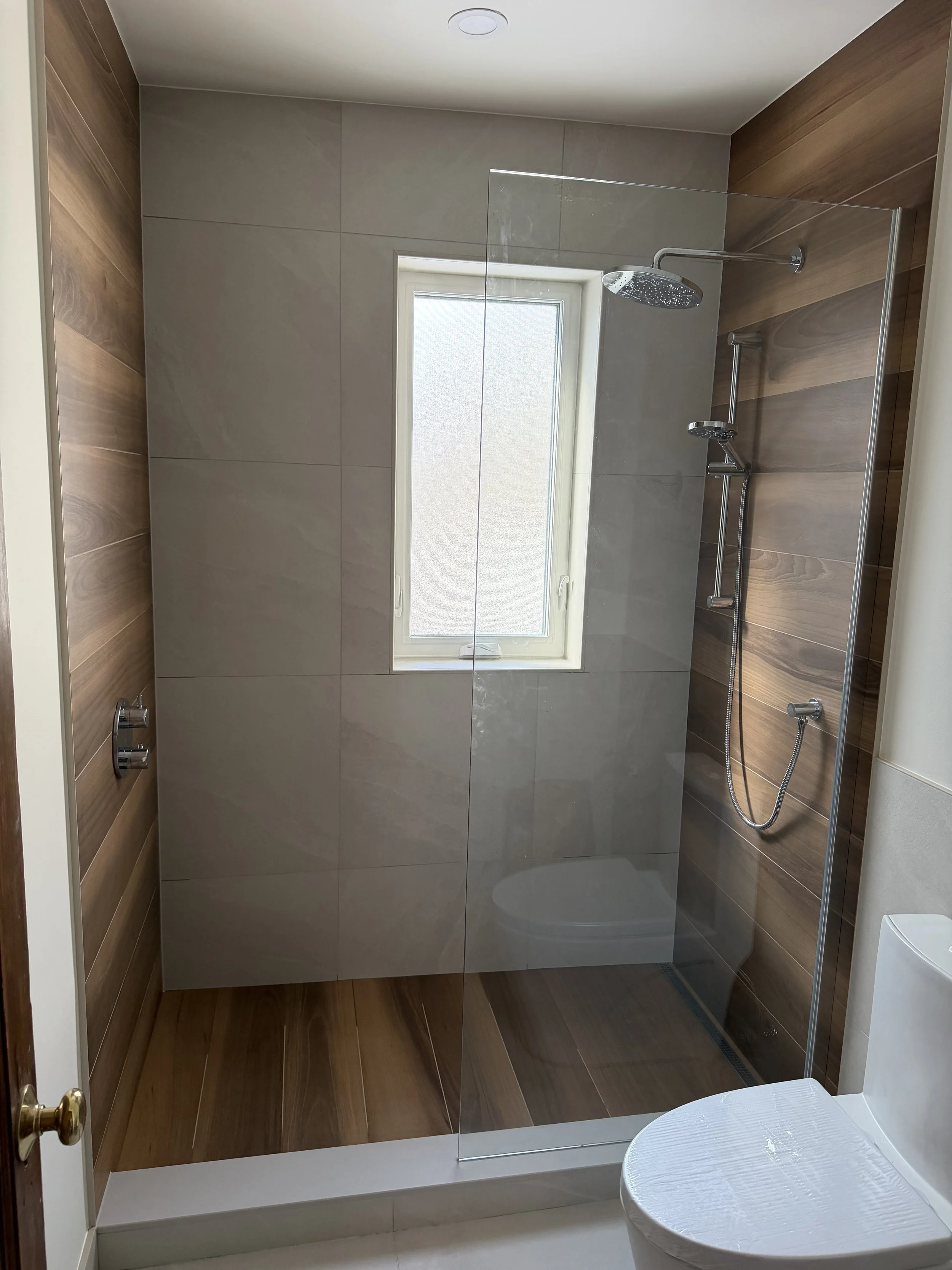 A modern bathroom with a walk-in shower featuring two shower heads and a glass divider. There is a small window on the back wall and the walls are decorated with large beige tiles and wood-like panels.
