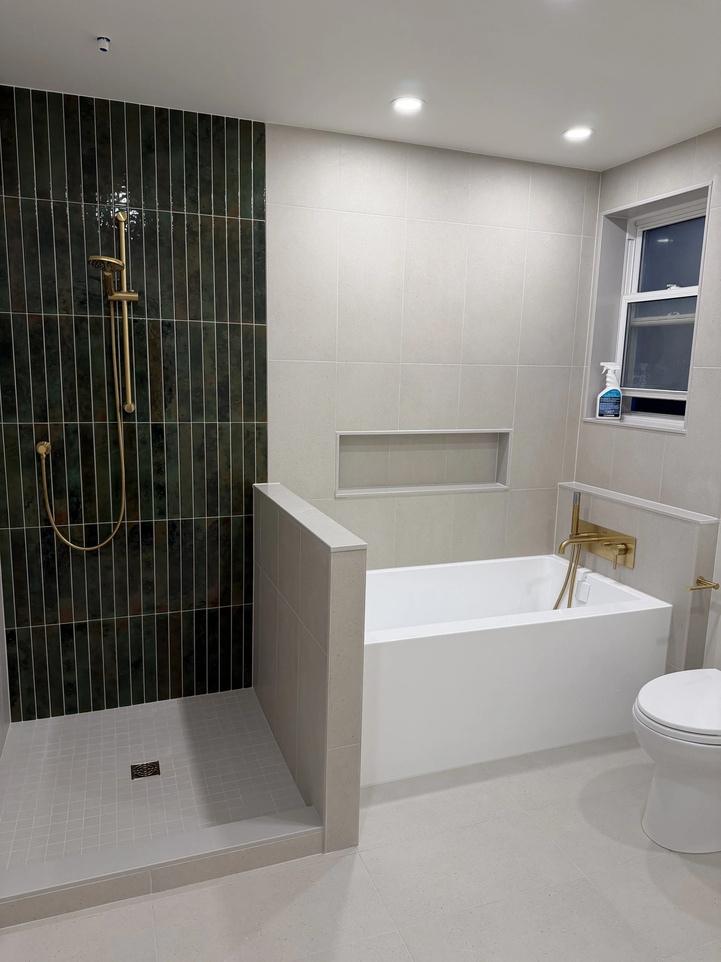 Modern bathroom with walk-in shower featuring dark green tile, white bathtub with gold fixtures, white toilet, and a window with cleaning spray.