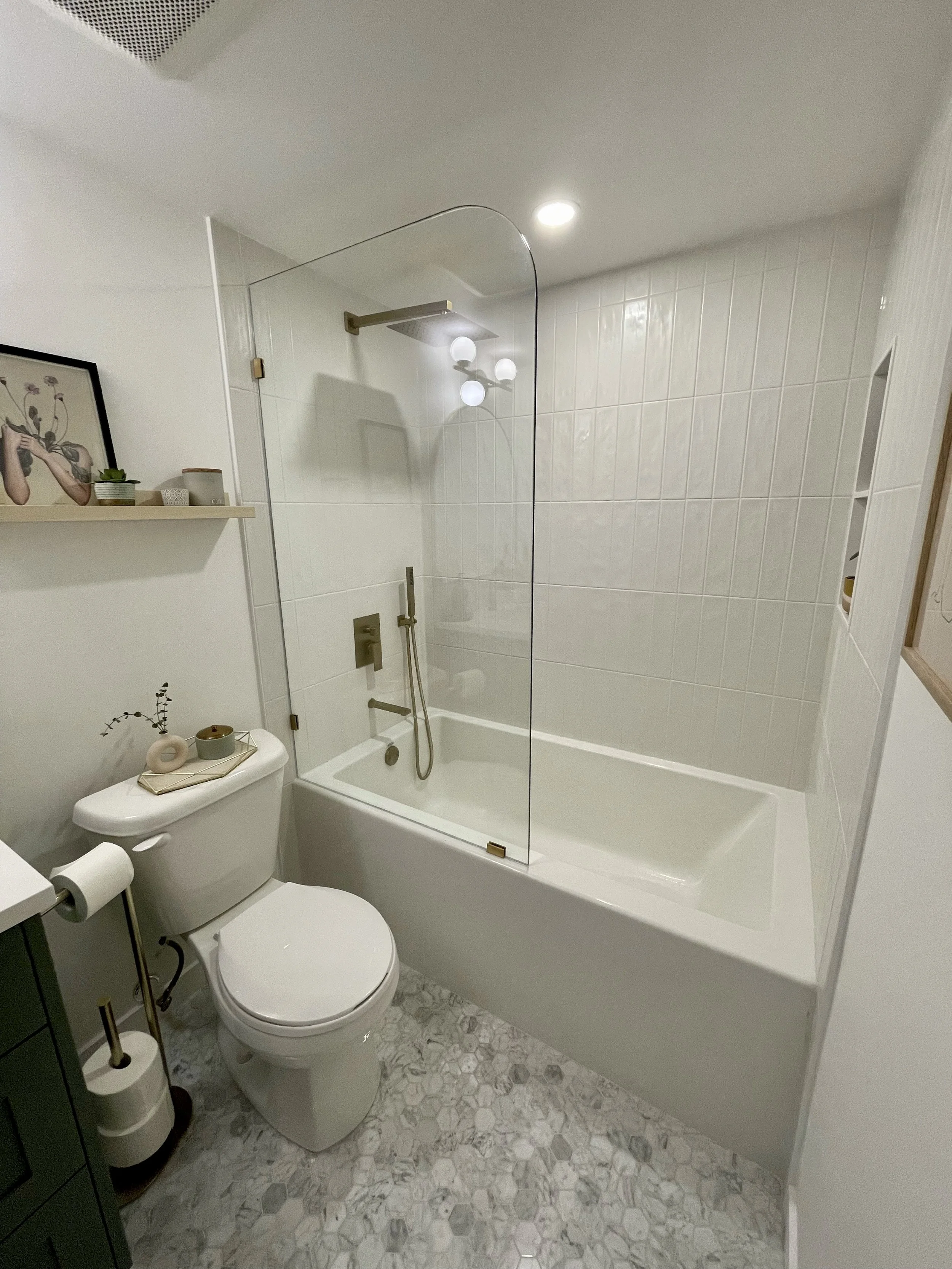 Modern bathroom with white bathtub, glass shower partition, white toilet, patterned tile floor, and minimal decor with small plants and artwork.