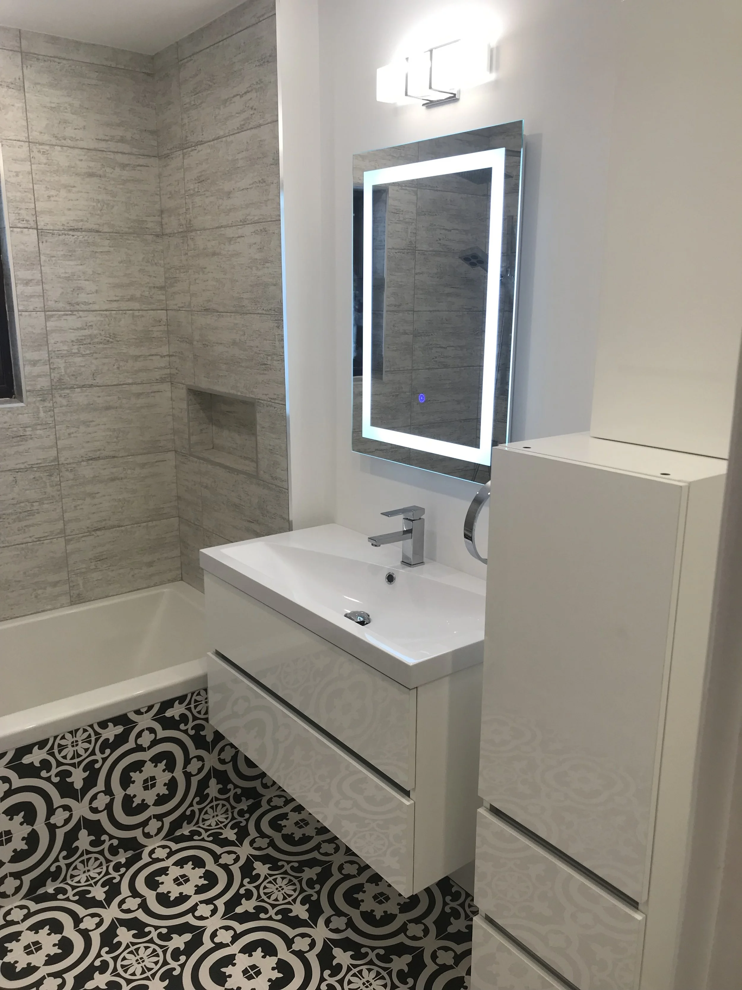 Modern bathroom with a white vanity, illuminated mirror, patterned black and white tile floor, beige tiled shower with built-in shelf, and white storage cabinet.