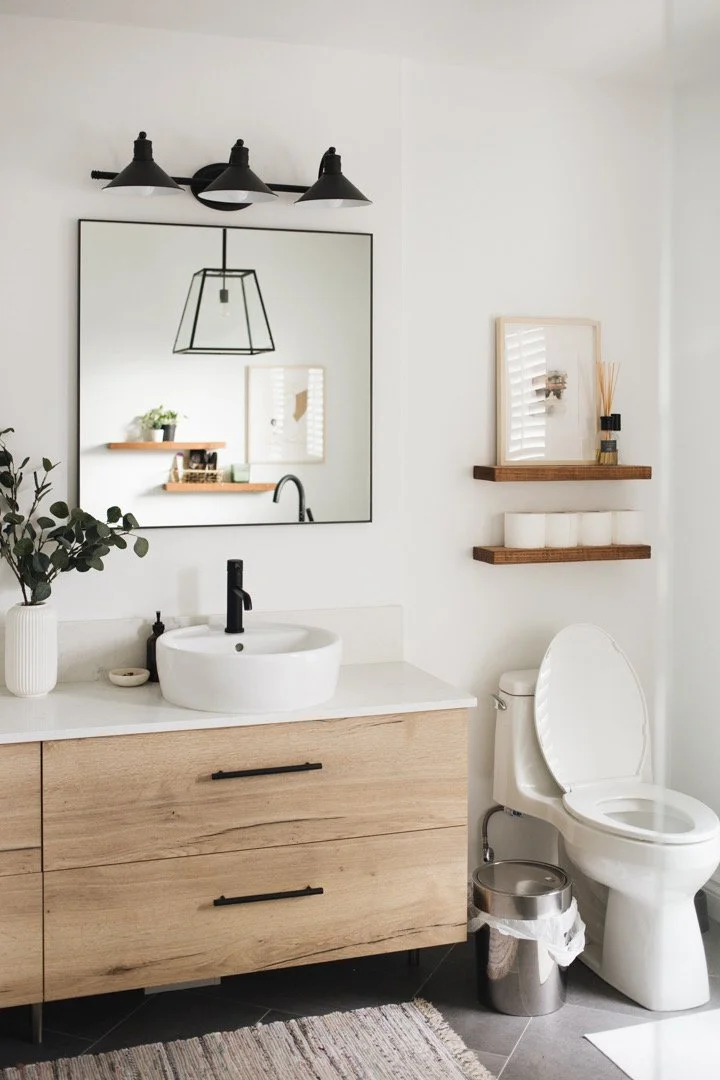 Modern bathroom with wooden vanity, white sink, black faucet, large mirror, wall-mounted light fixture, potted plant, floating shelves with decor, toilet, and trash bin.