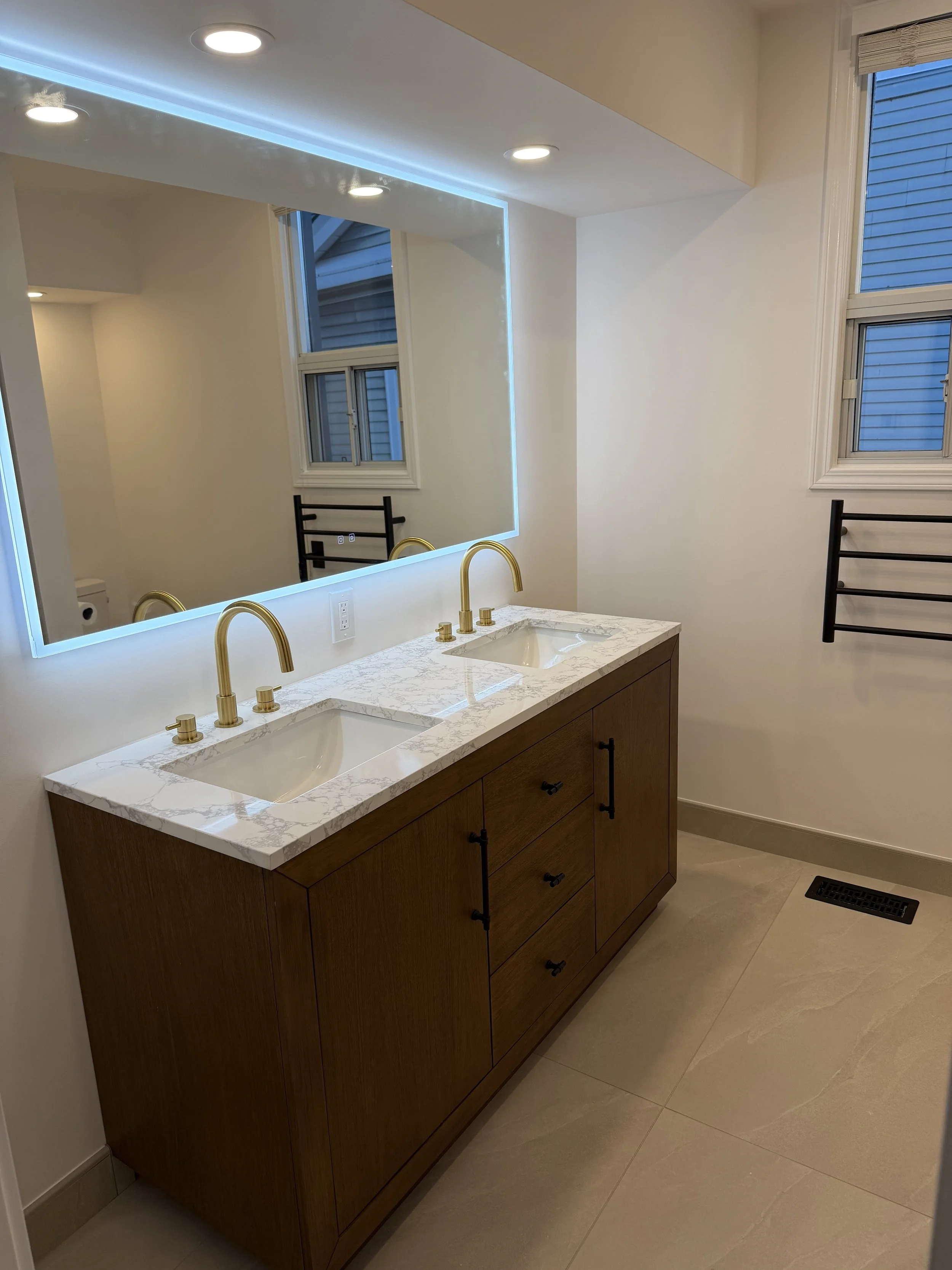Bathroom vanity with dual sinks, marble countertop, gold faucets, large backlit mirror, window with blinds, and heated towel racks.