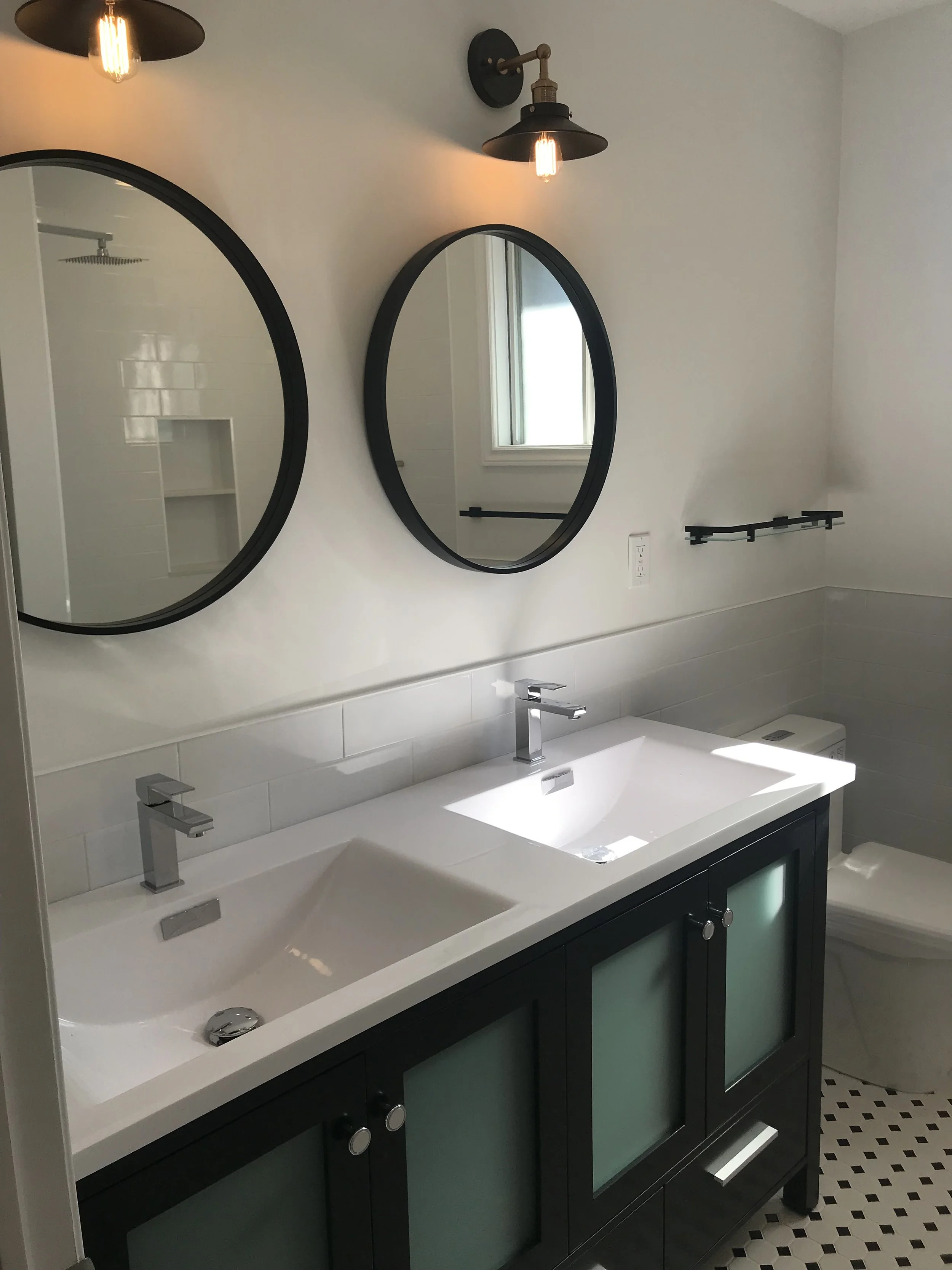 A modern bathroom with a dual vanity sink with black cabinets and frosted glass doors, two oval mirrors, black wall-mounted lamps, a small window, and a shower with a built-in shelf area.