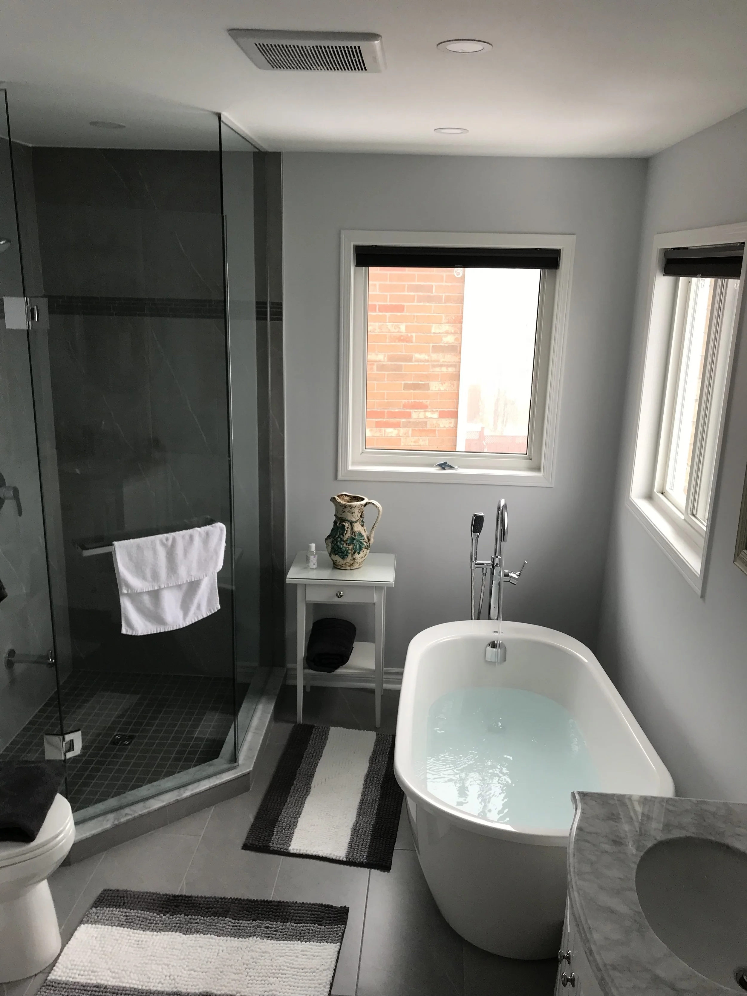 Modern bathroom with a white bathtub, a shower with glass doors, and a small white table with a decorative pitcher. There are two windows letting in natural light, and the bathroom is decorated in neutral tones with striped rugs on the floor.
