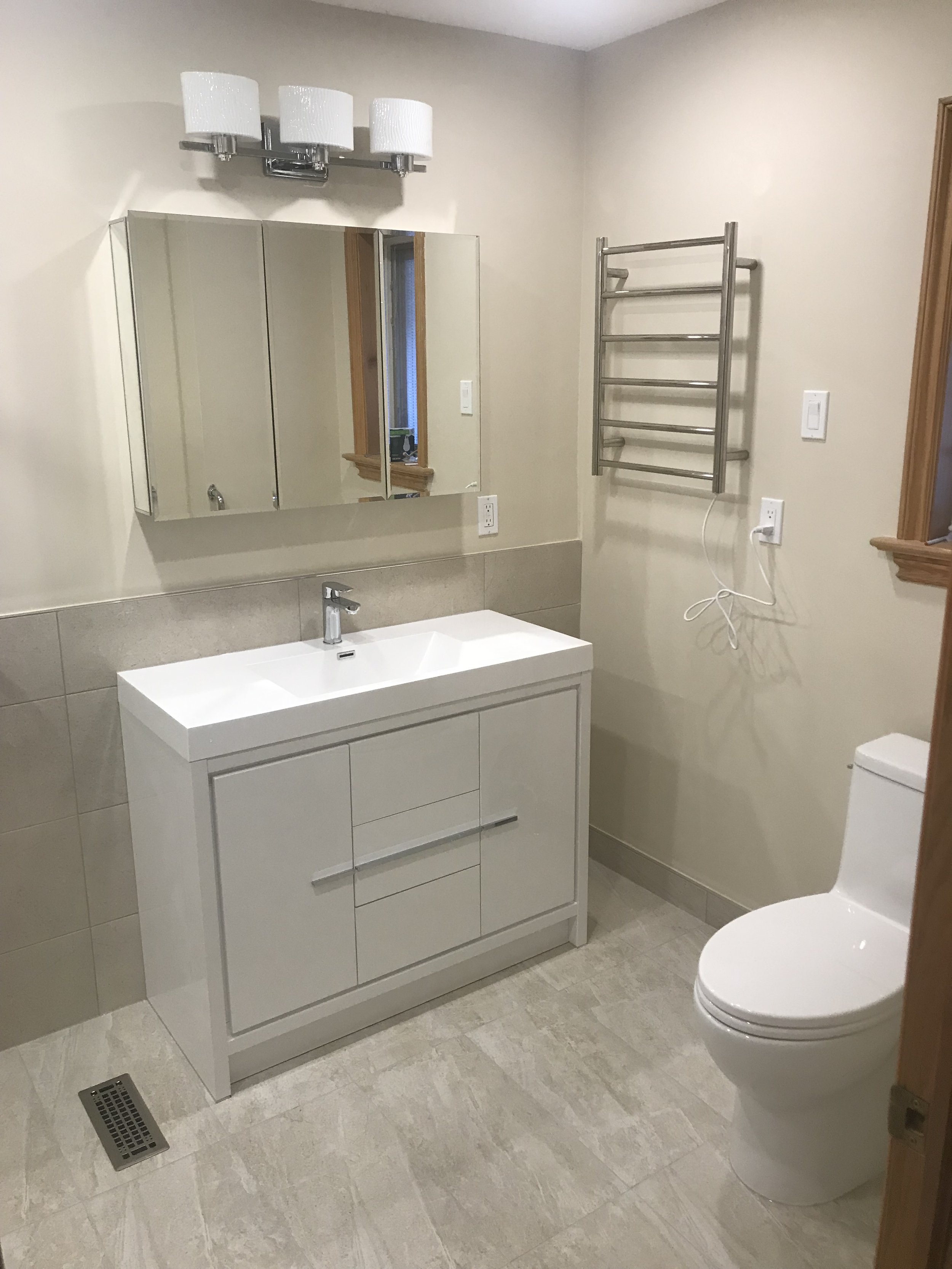 Bathroom with a white vanity, mirror cabinet, wall-mounted towel warmer, and a toilet. The vanity has a sink with a faucet and three drawers. The mirror cabinet has three mirrored doors. A light fixture with four lights is mounted above the mirror. T