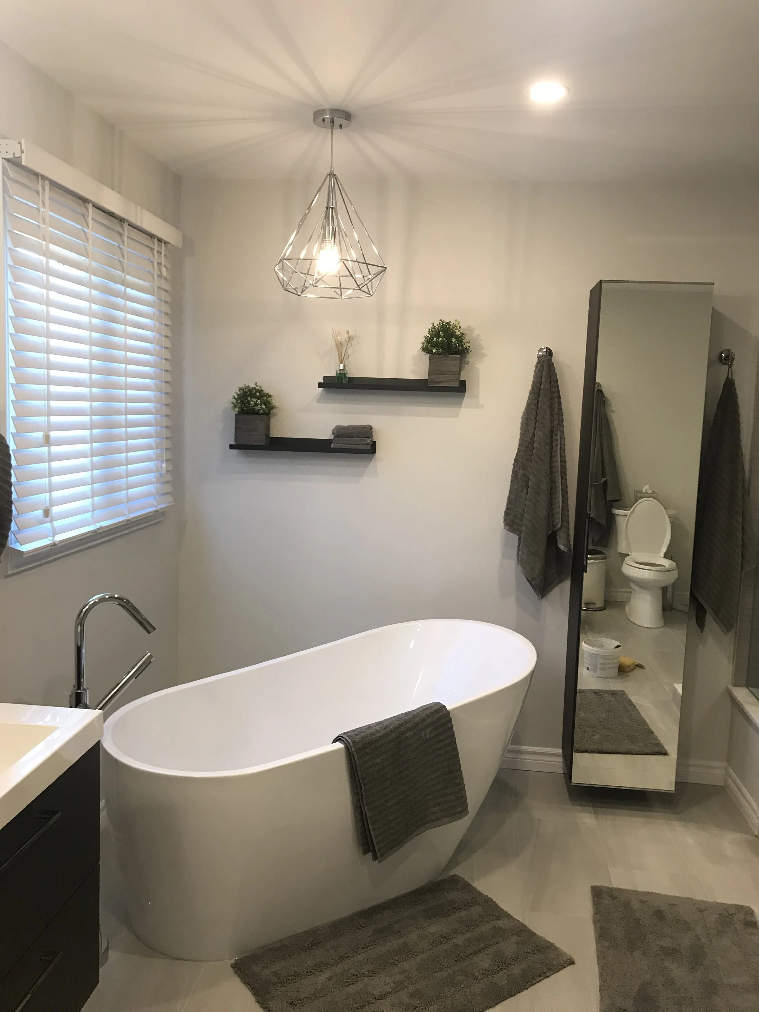 Modern bathroom with a white freestanding bathtub, gray towels, a large mirror, wall-mounted black shelves with small potted plants, a gray bathroom mat, a toilet in the background, and a geometric ceiling light fixture.