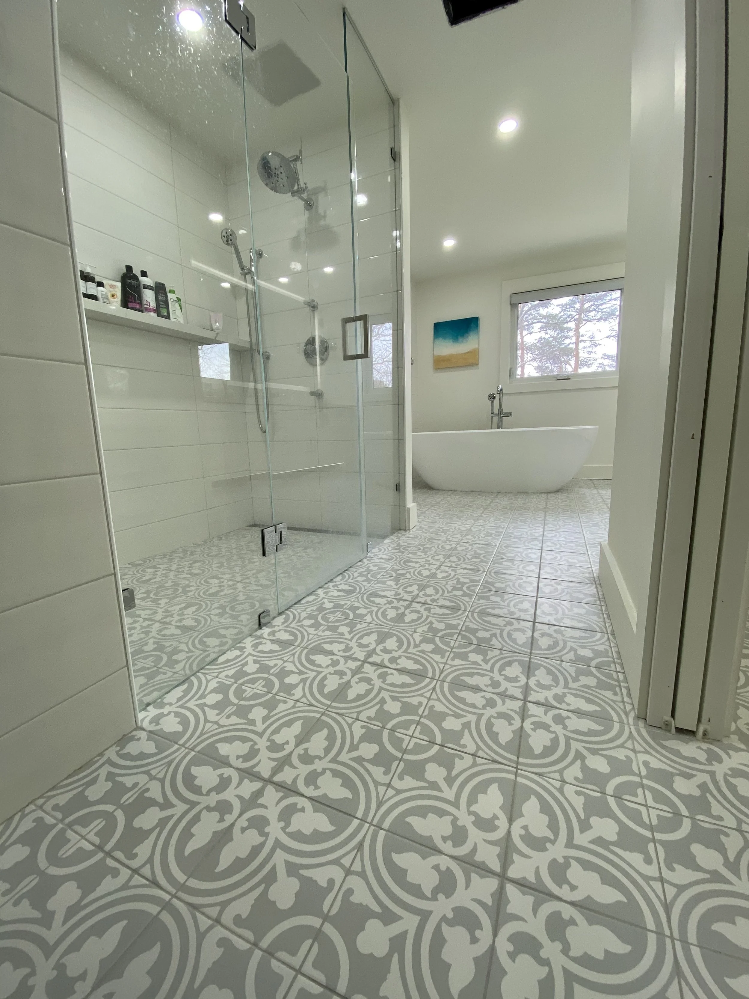 Bright bathroom with patterned floor tiles, a glass-enclosed shower stall, a free-standing bathtub, a window with trees outside, and wall art.