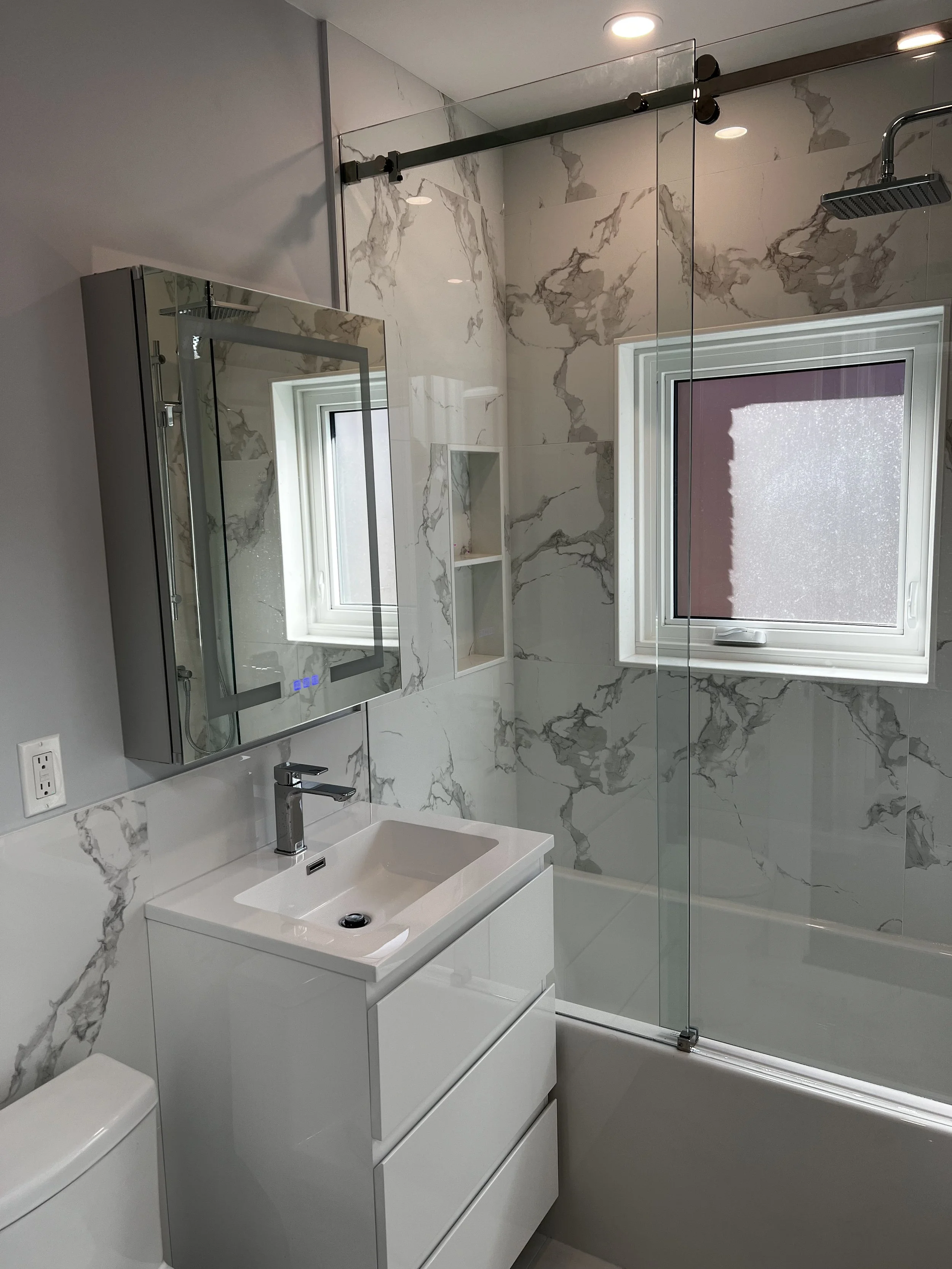 Modern bathroom with marble-patterned wall tiles, a rectangular white sink with a cabinet, a mirror with integrated lighting, a window with frosted glass, and a glass shower enclosure with a rainfall showerhead.