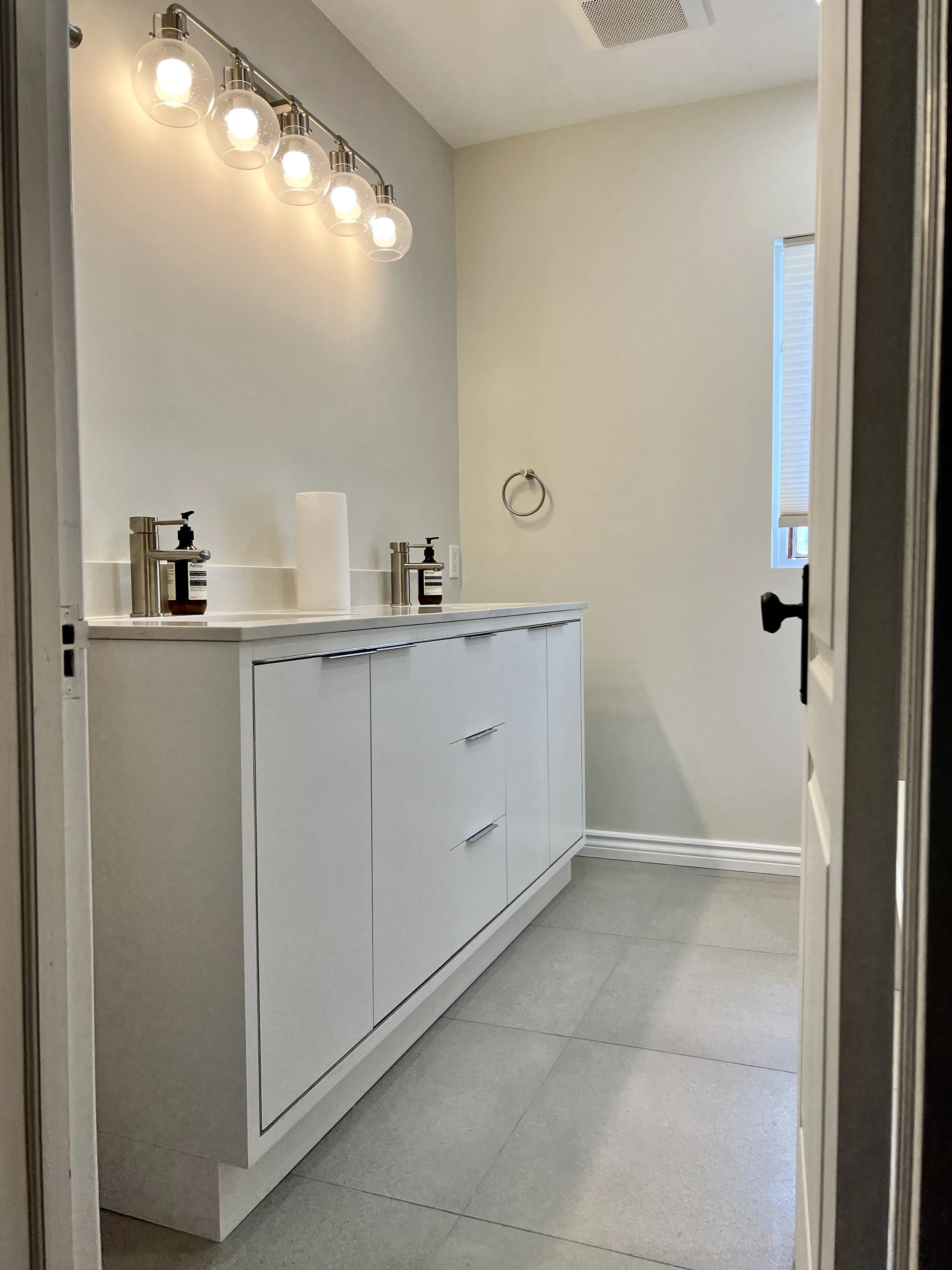 A modern bathroom with a white vanity, two sinks with stainless steel faucets, soap dispensers, a paper towel roll, a towel ring, a window with a blind, and a row of six globe lights above the mirror.
