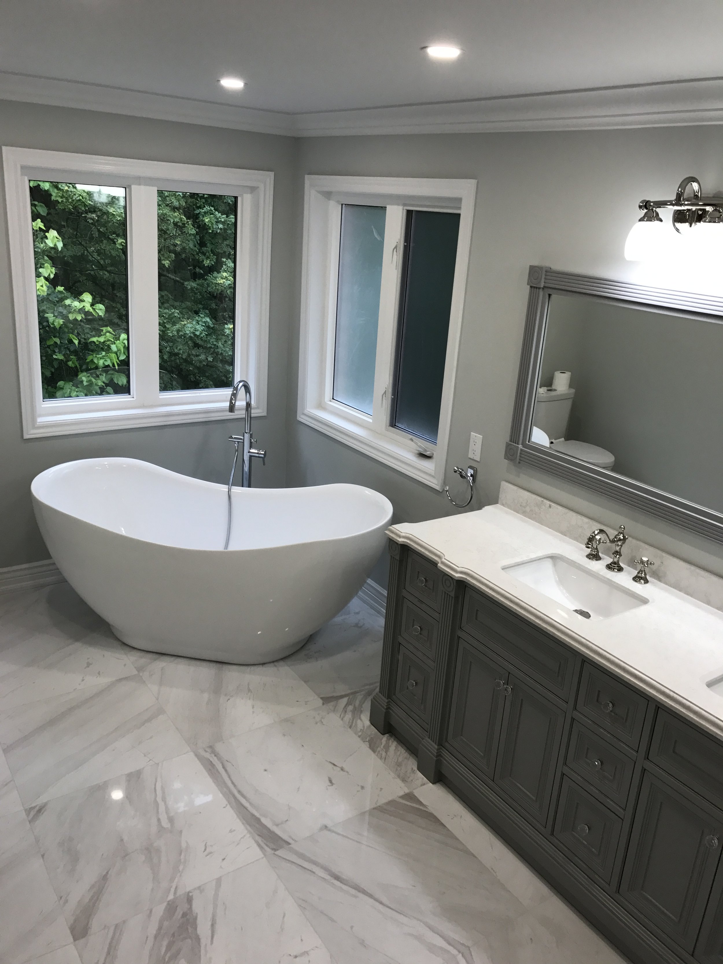 Modern bathroom with a freestanding bathtub, gray vanity with a sink, large mirror, marble floor, windows showing green trees, and a toilet in the background.