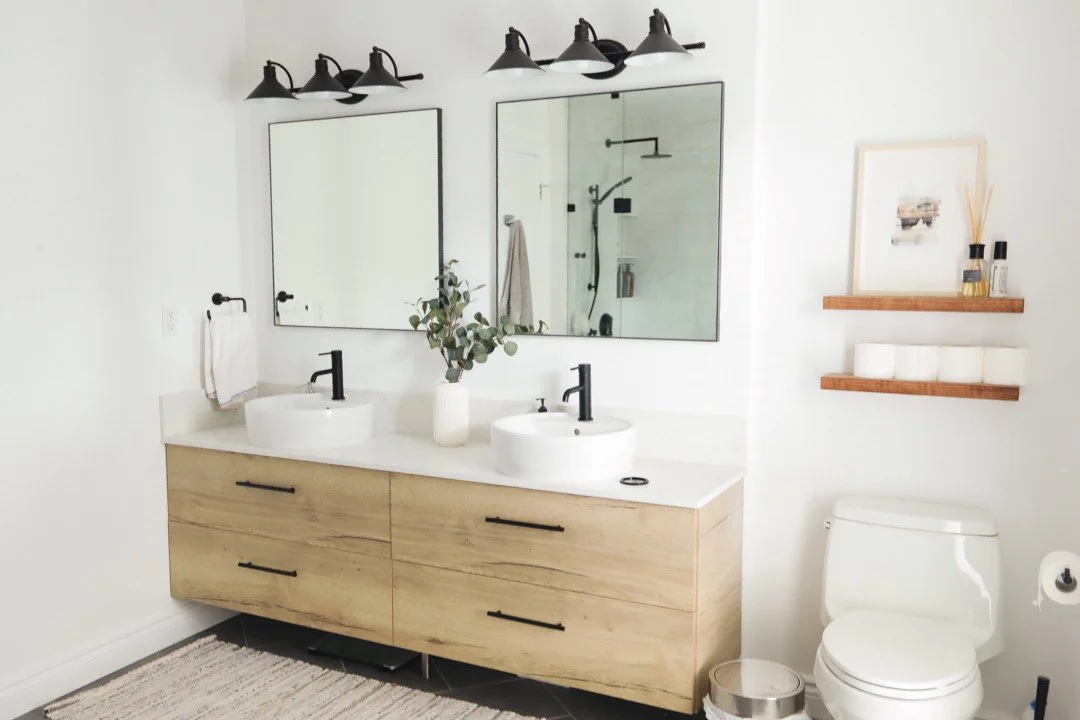 Modern bathroom with a wooden vanity, two white vessel sinks with black faucets, two large mirrors, and black fixture lights above. There is a small plant in a white vase, a towel, a framed picture, and shelves with rolled towels and toiletries. A to