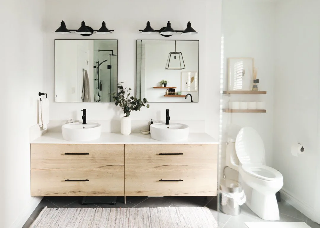 Modern bathroom with double vanity, two round vessel sinks, black faucets, two mirrors, black light fixtures, a toilet, and wooden shelves with decor.
