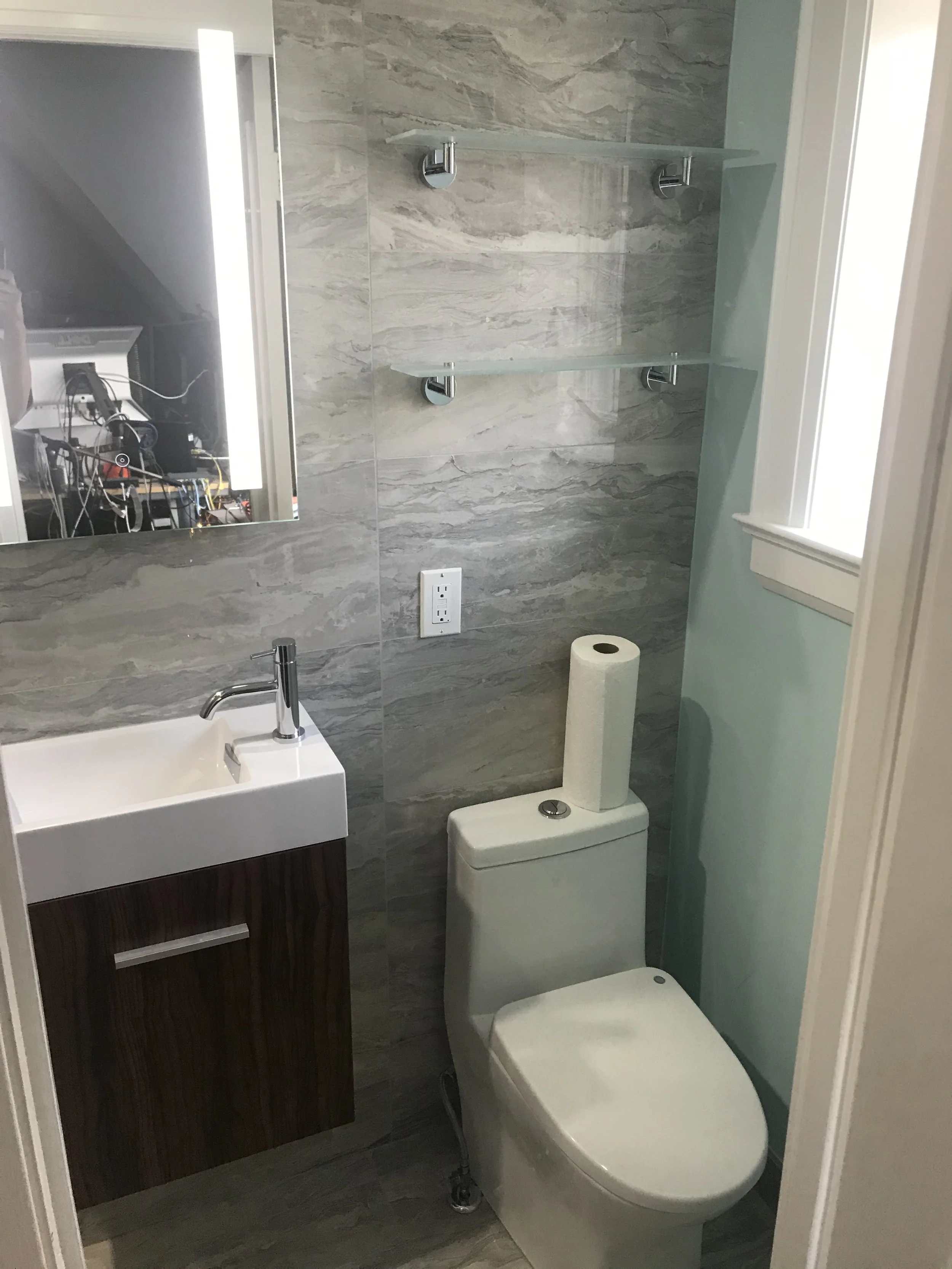 A small bathroom with a wall-mounted mirror, a white sink cabinet, a modern toilet with a roll of toilet paper on top, two glass shelves on the wall, a window, and an electrical outlet with a cover. The bathroom features gray marble-patterned wall ti