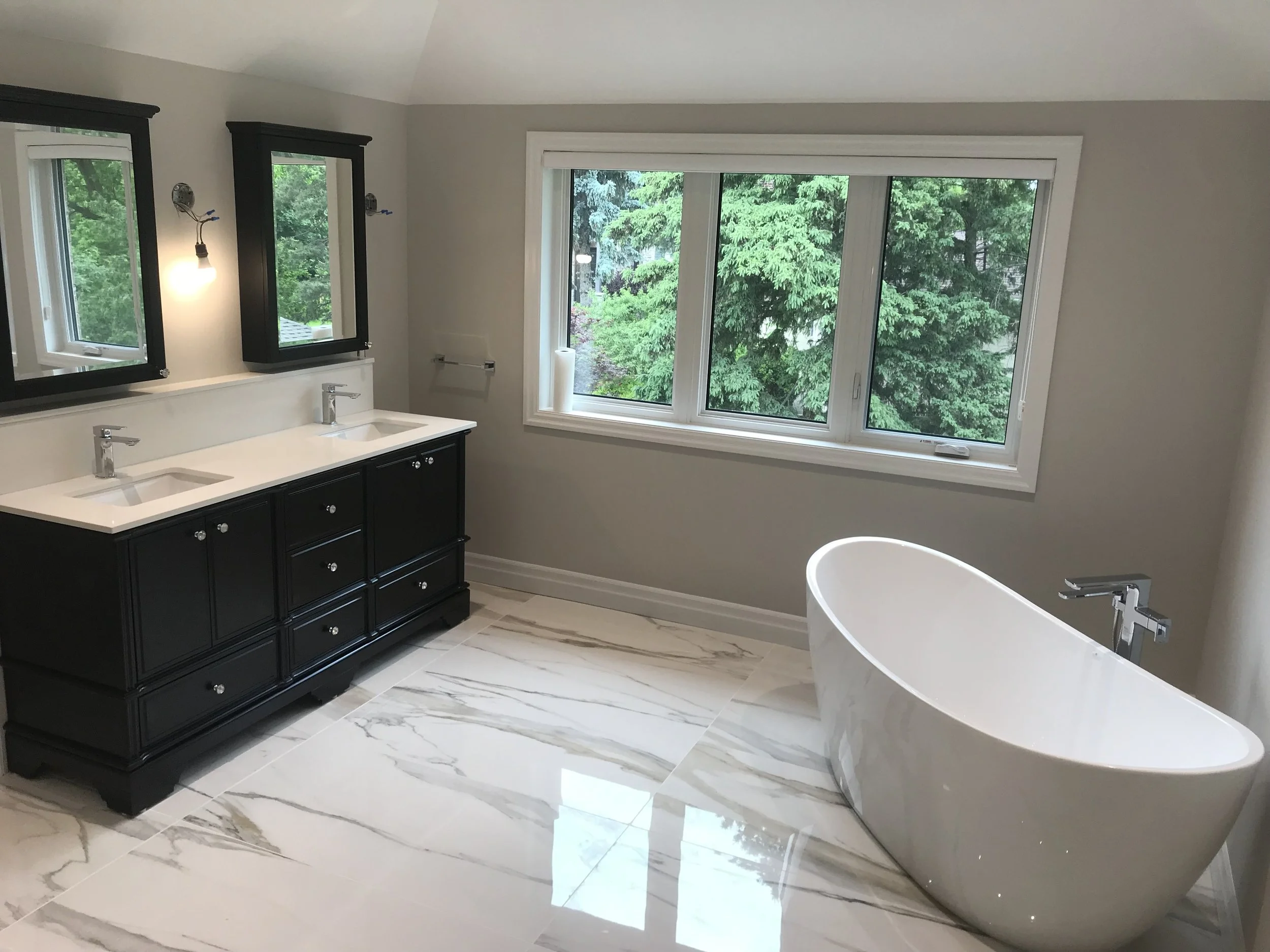 Modern bathroom with a freestanding bathtub, a dual-sink vanity with black cabinets, and a large window overlooking green trees.