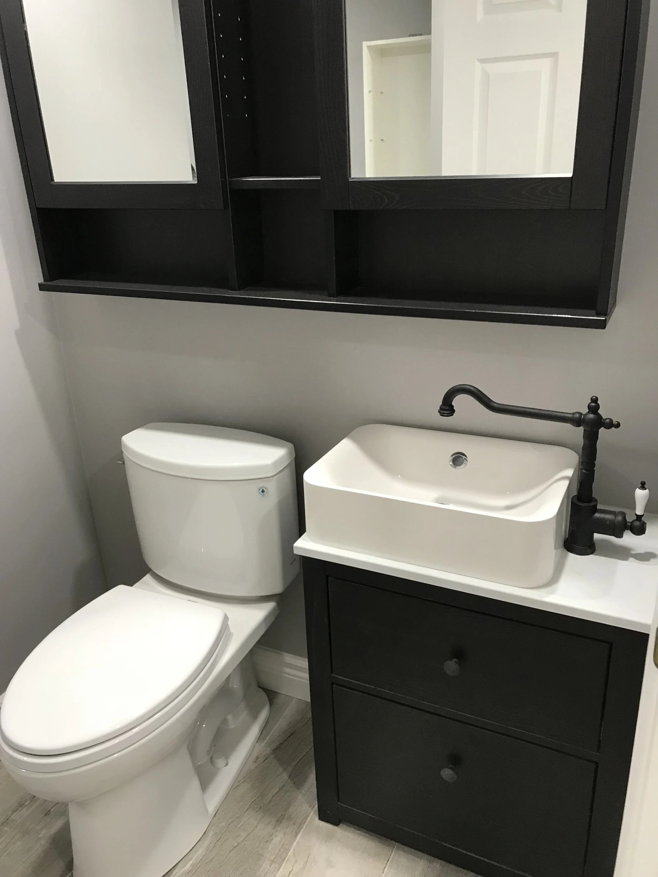 A small bathroom with a white toilet, a black vanity with two drawers, a rectangular white vessel sink, a black faucet, and a black wall-mounted cabinet with mirrored doors.