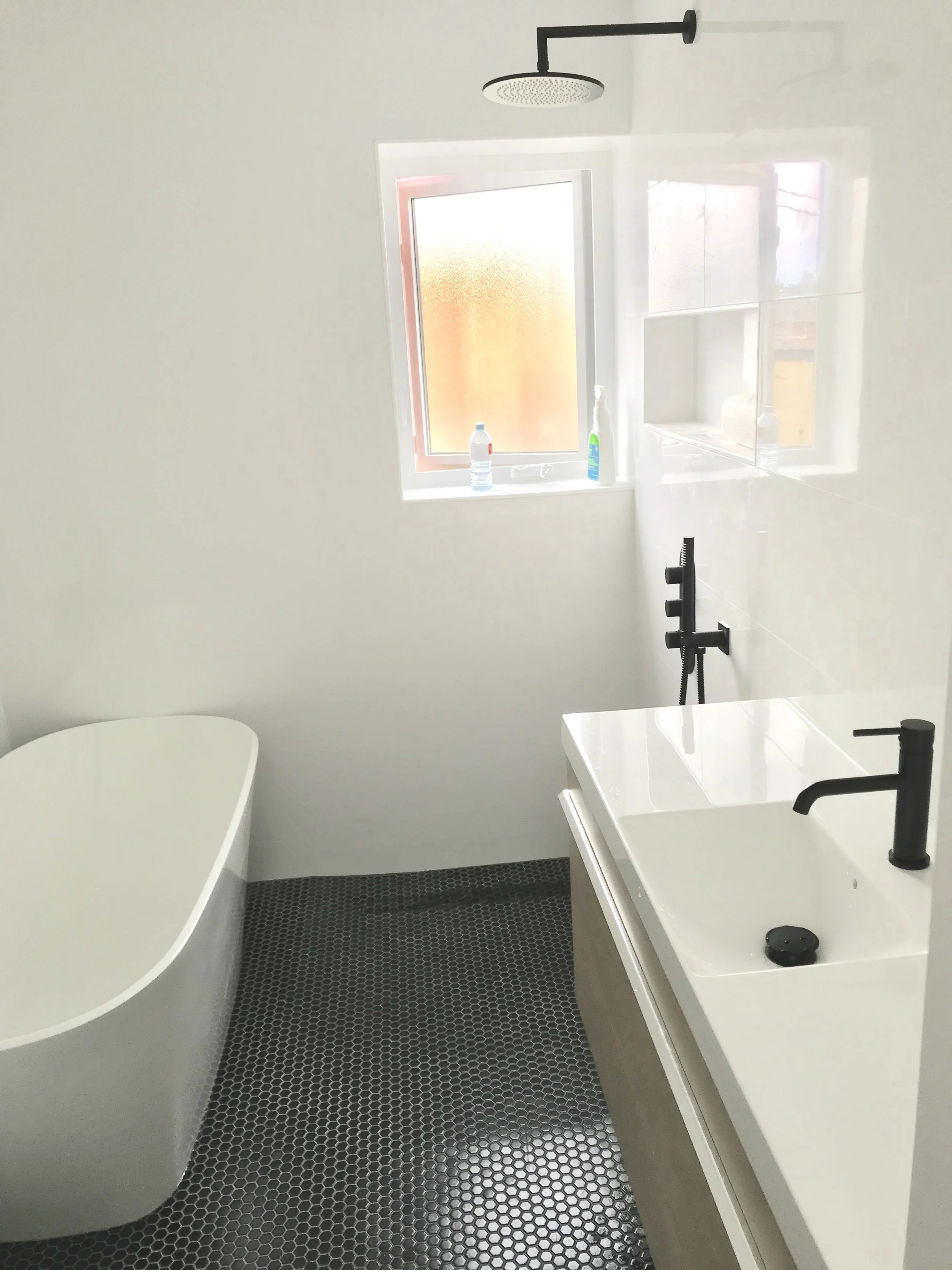 Modern bathroom with white bathtub, black matte fixtures, small black hexagon tile floor, white walls, and a window with cleaning supplies on the sill.