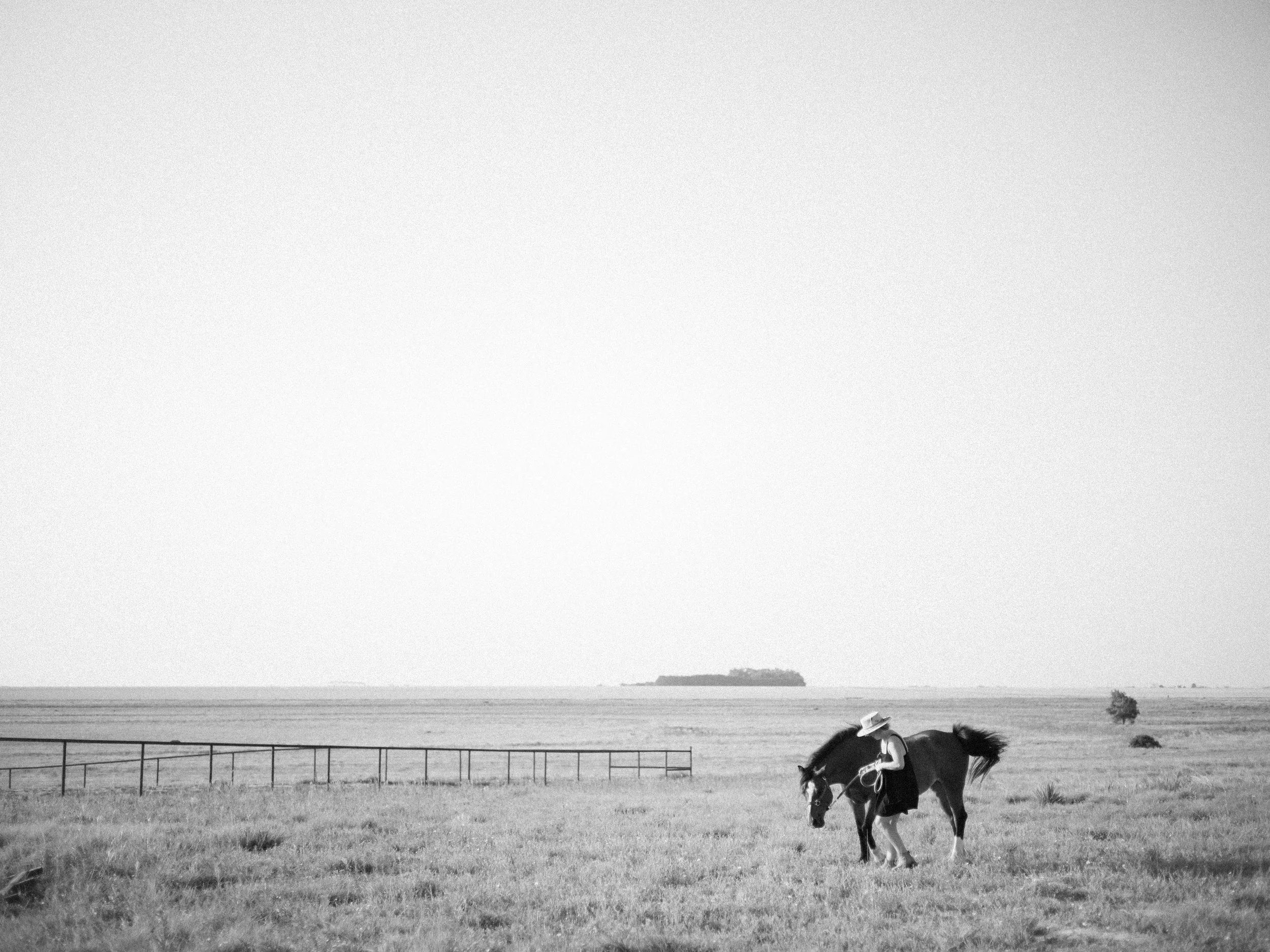 A person wearing a hat and dress, sitting on a horse in a wide open grassy field with a fence in the distance and a tree, with a large expanse of sky above.