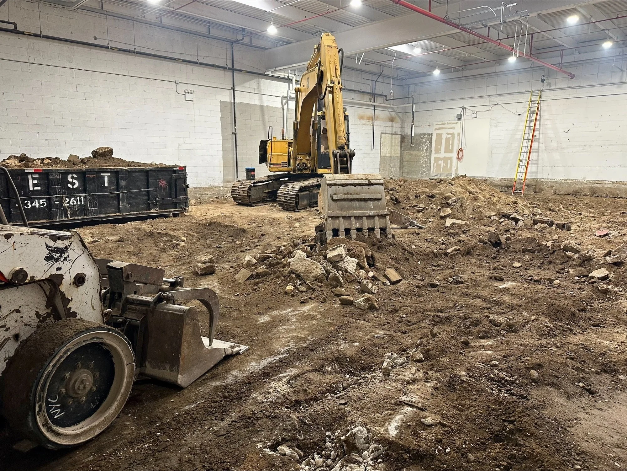 Construction site inside a warehouse with a yellow excavator, rubble, a roller compactor, and a ladder against the wall.