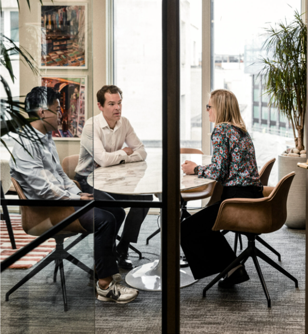 Three people sitting at a round table in a modern office meeting room with large windows, indoor plants, and colorful artwork on the wall, engaged in a discussion.