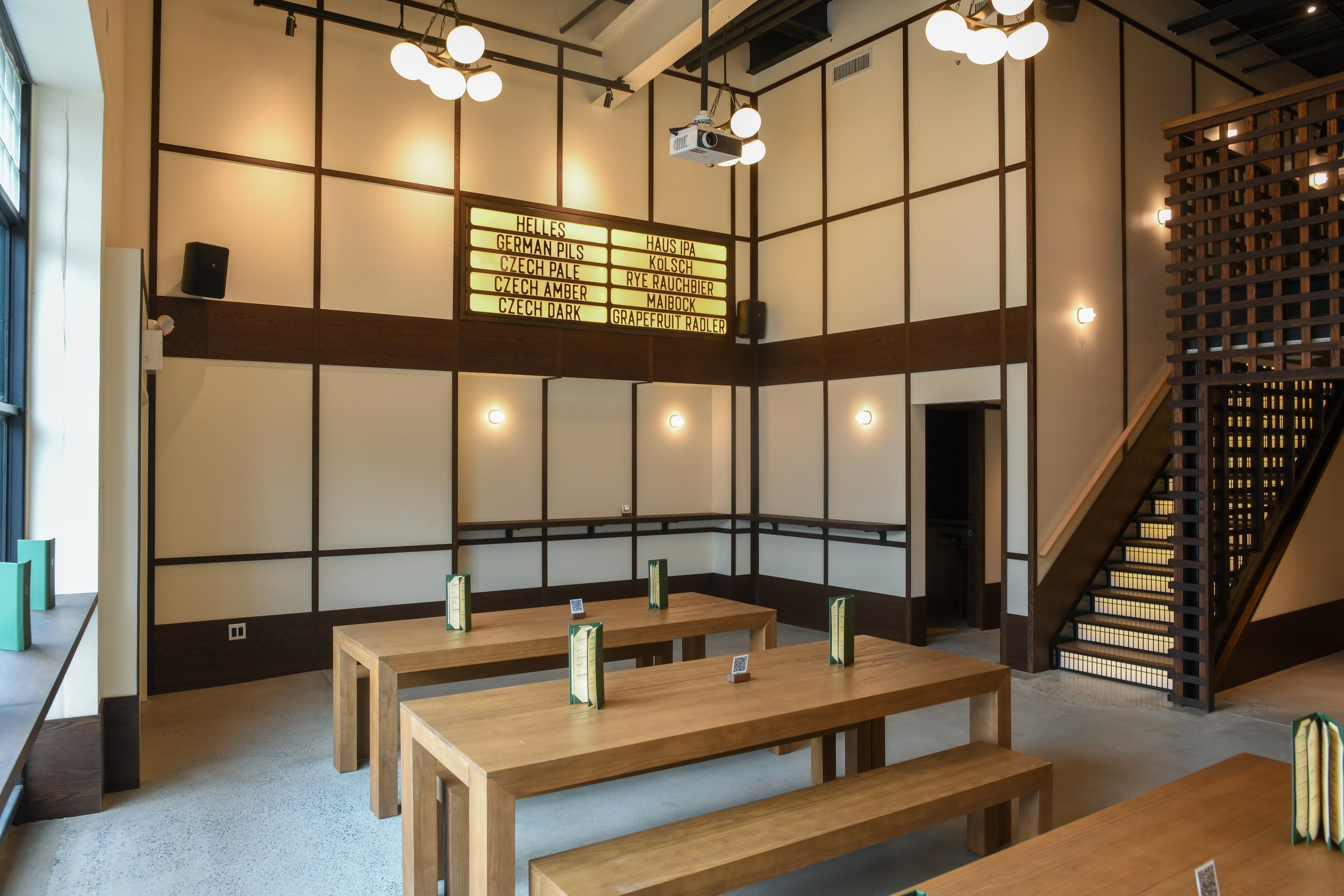 A modern interior space with wooden tables and benches, beige walls with dark wooden paneling, hanging ceiling lights, a staircase on the right, and a yellow-lit beer menu board on the back wall.