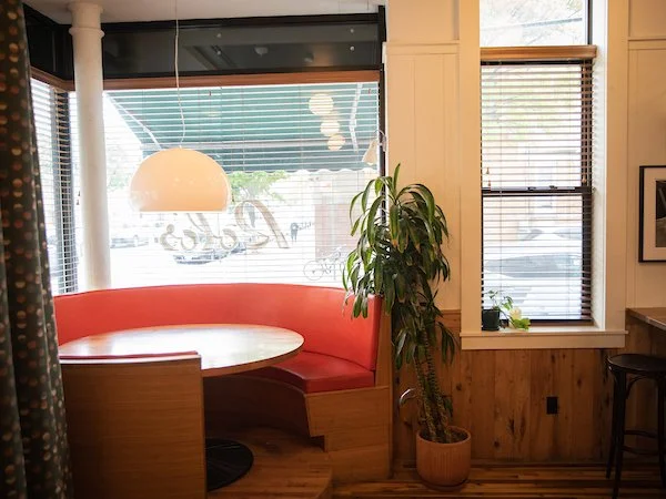 Corner booth with red cushioned seating, wooden table, large potted plant, window with blinds, and a hanging light fixture inside a cozy cafe.