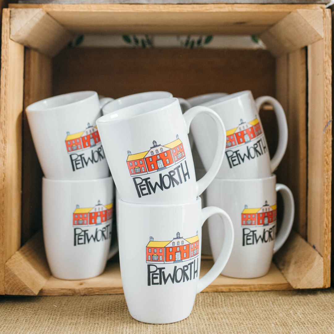 White coffee mugs with a colorful logo of a red building and the word PETWORTH stacked on top of each other in a wooden crate.