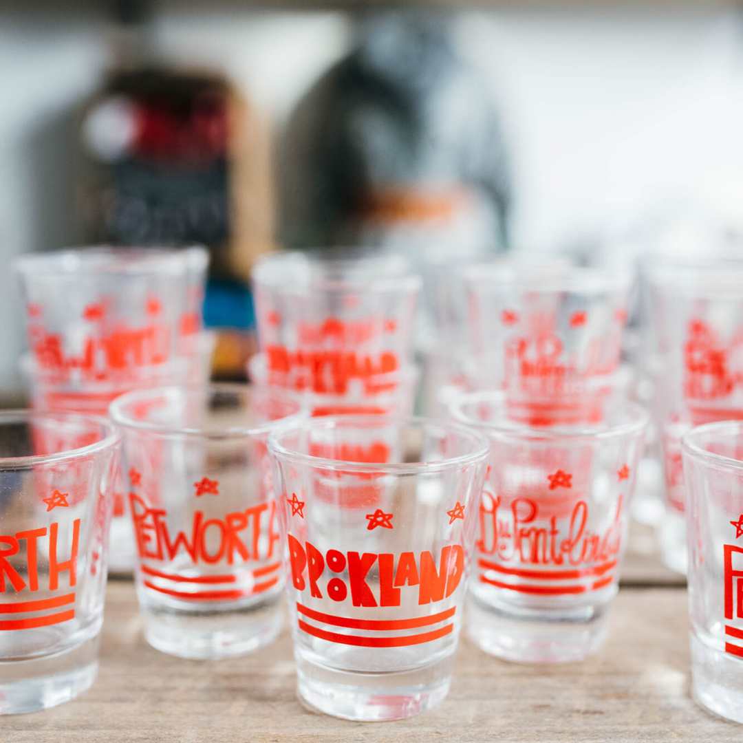 Clear glass cups with red text and stars, labeled with different city names, arranged on a wooden surface in a row.