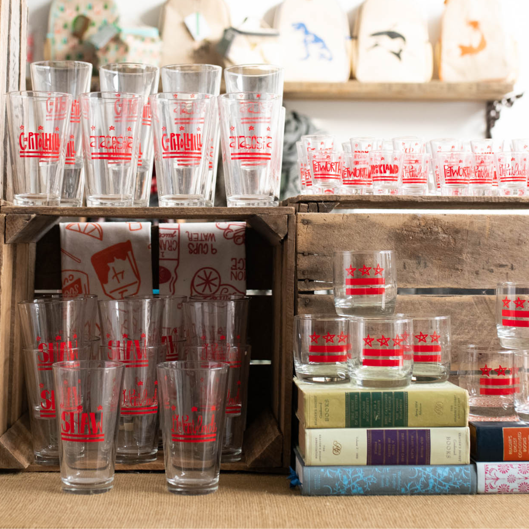 Display of vintage glassware with red patriotic designs, books, and decorative tote bags on a wooden shelf in a store.