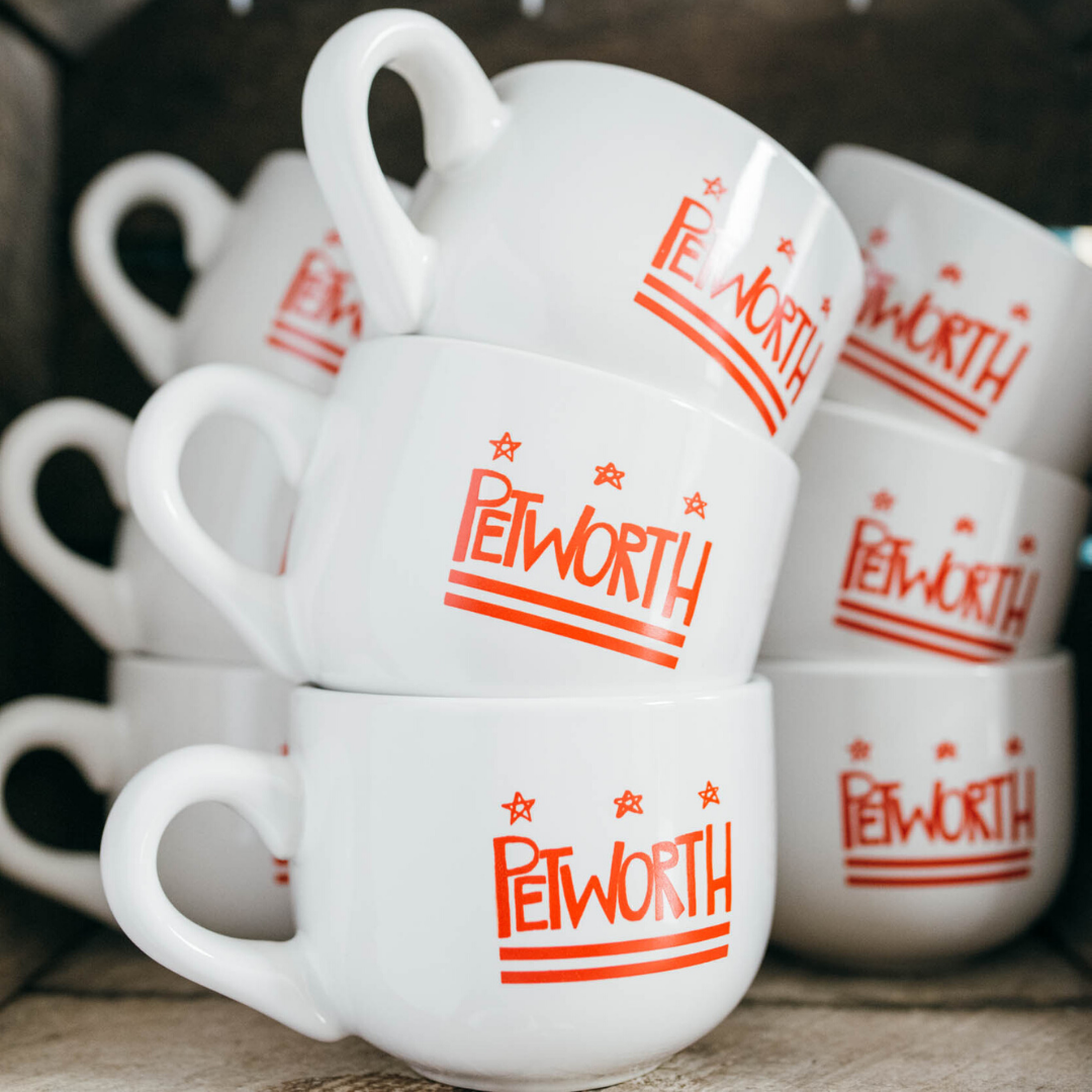 Stack of white ceramic coffee mugs with red PetWorth logo and stars, arranged on a wooden surface.