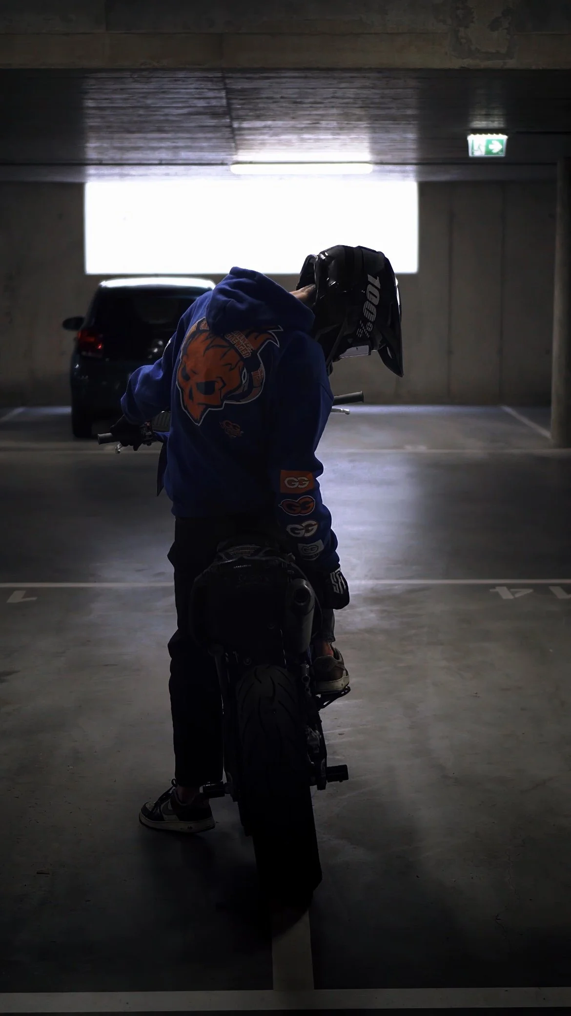 Junge in dunkler Helmmütze und blauer Hoodie mit auffälligem Logo auf dem Rücken, steht in einer Tiefgarage neben einem Motorrad, bei Gegenlicht. Ein Auto ist im Hintergrund zu sehen.