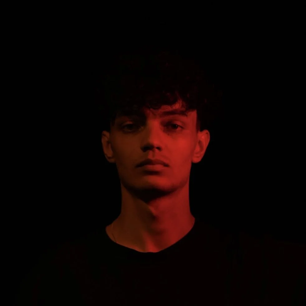 Porträt eines jungen Mannes mit dunklen Haaren und dunkler Kleidung vor schwarzem Hintergrund, beleuchtet mit rotem Licht.