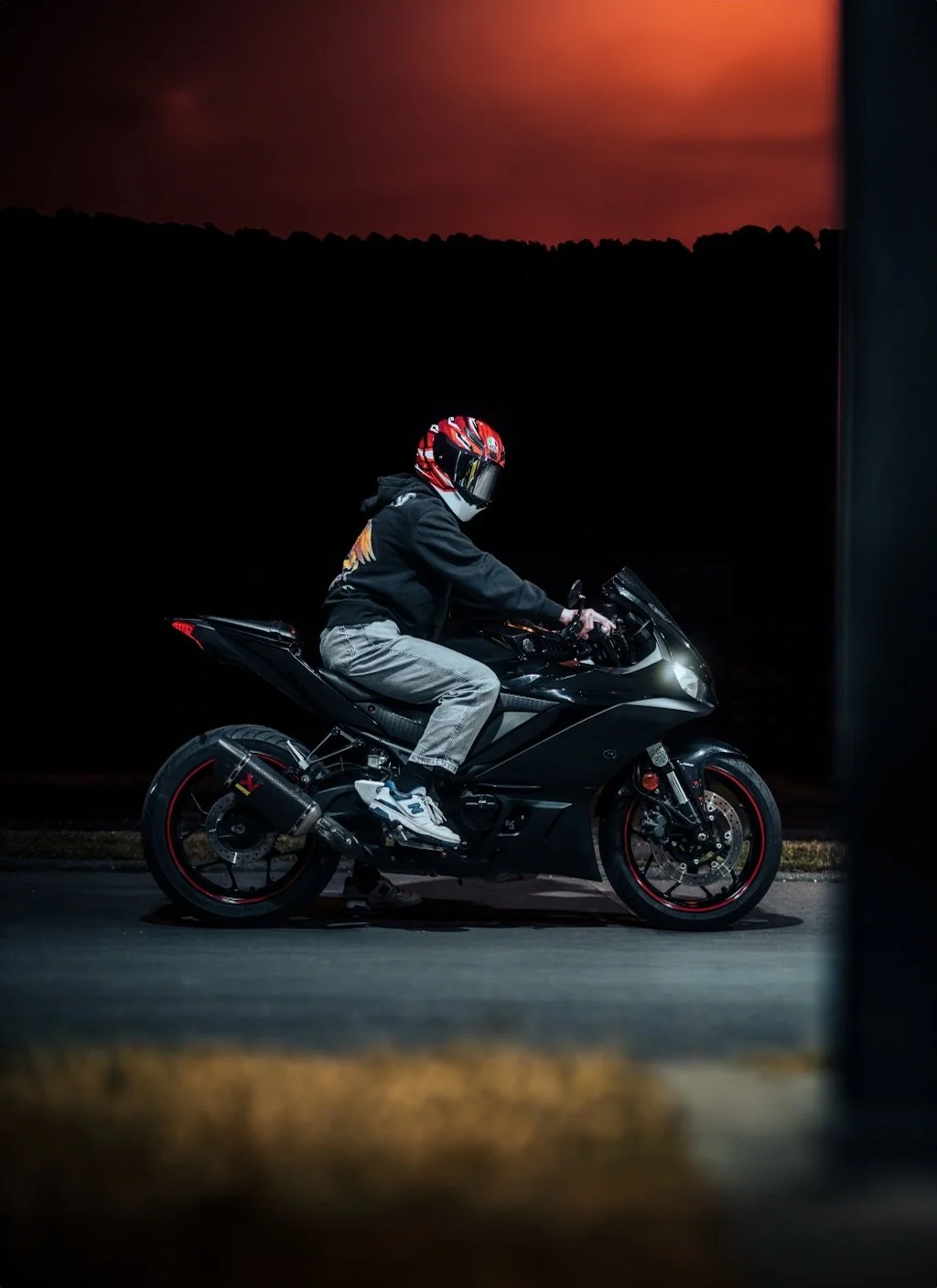 Ein Motorradfahrer in schwarzer Jacke, grauen Hosen und rotem Helm sitzt auf einem schwarzen Sportmotorrad bei Nacht, im Hintergrund ein roter Himmel.