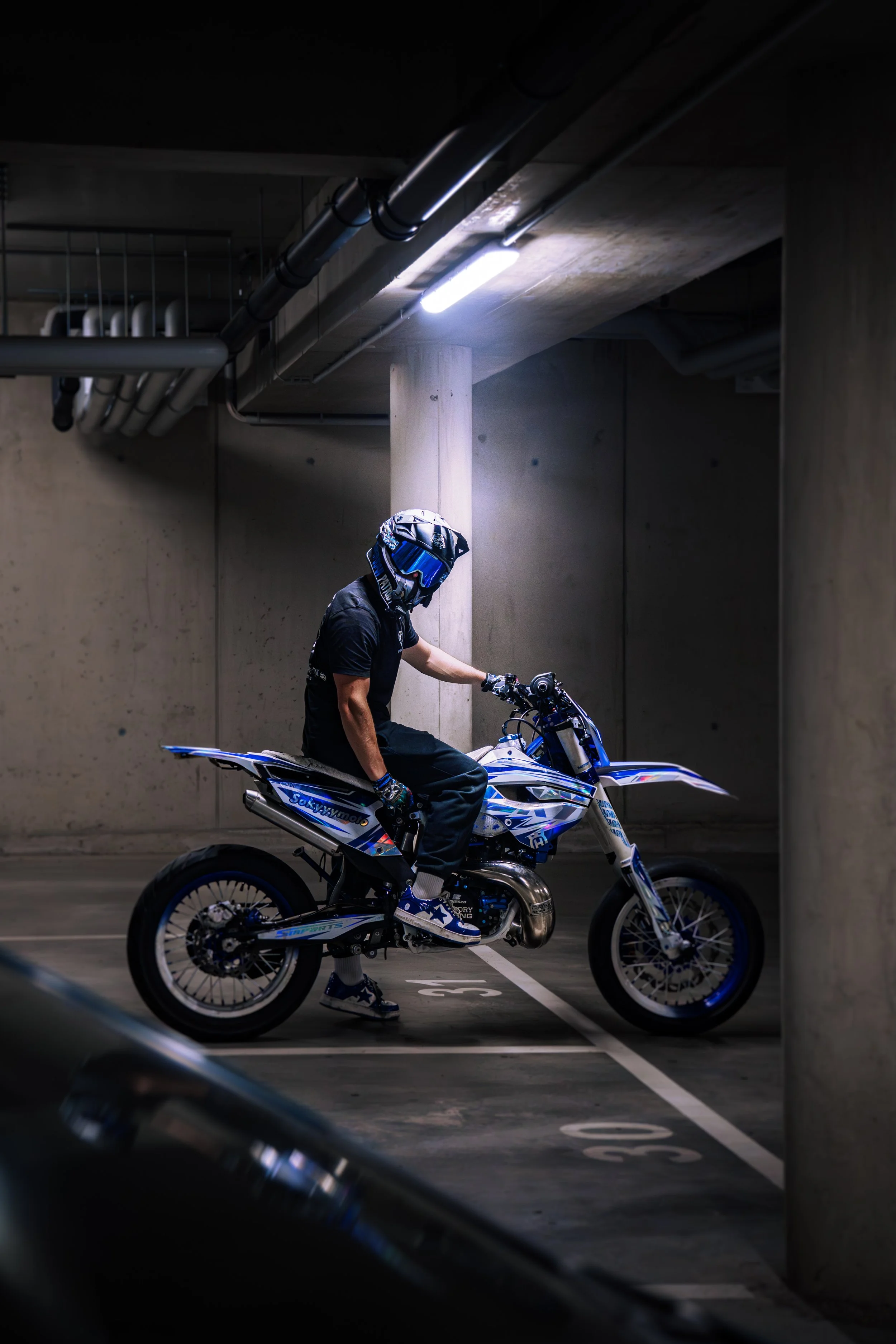 Ein Motorradfahrer in voller Schutzkleidung, einschließlich Helm und Handschuhe, sitzt auf einem blauen Offroad-Motorrad in einer Tiefgarage, die von Deckenbalken und Rohrleitungen überdacht ist.