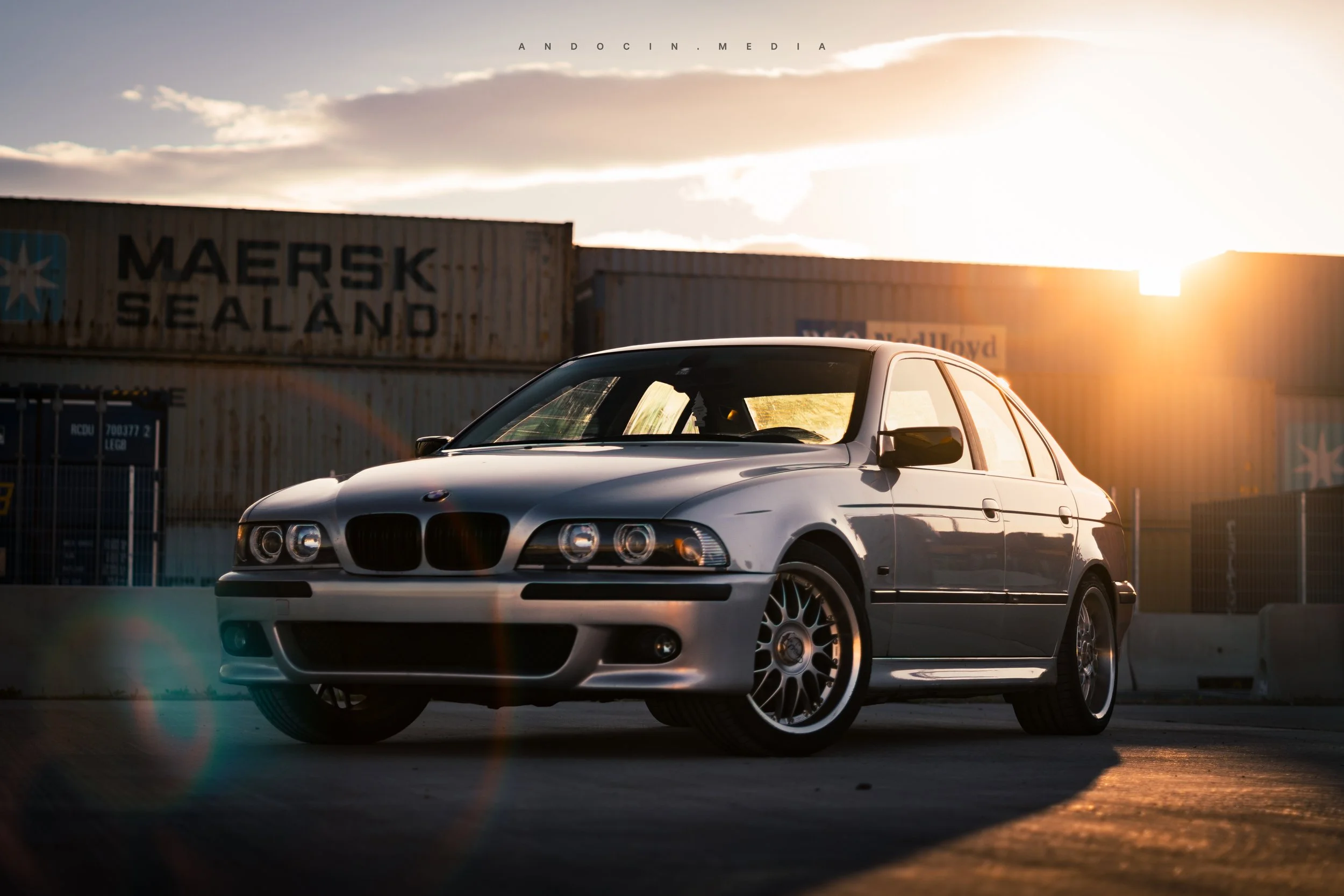 Ein silberner BMW-Einstiegswagen steht bei Sonnenuntergang vor Containern mit der Aufschrift 'Maersk Sealand'.