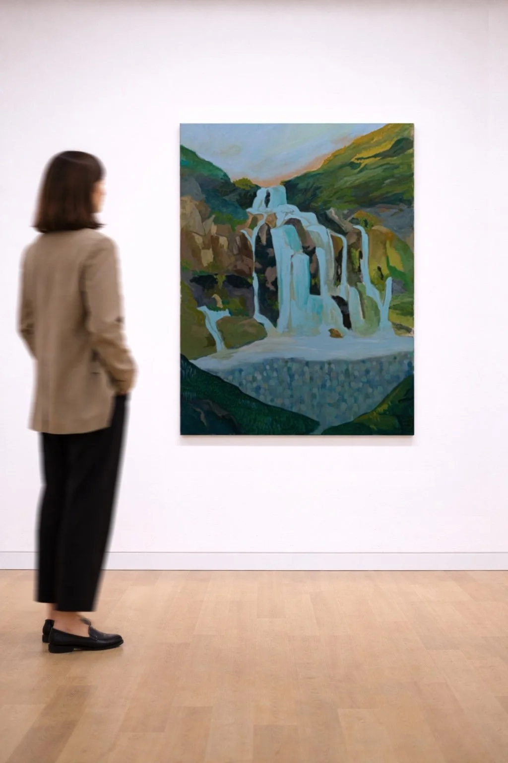 A woman observing a landscape painting of a waterfall surrounded by green hills in an art gallery with white walls and wooden floor.