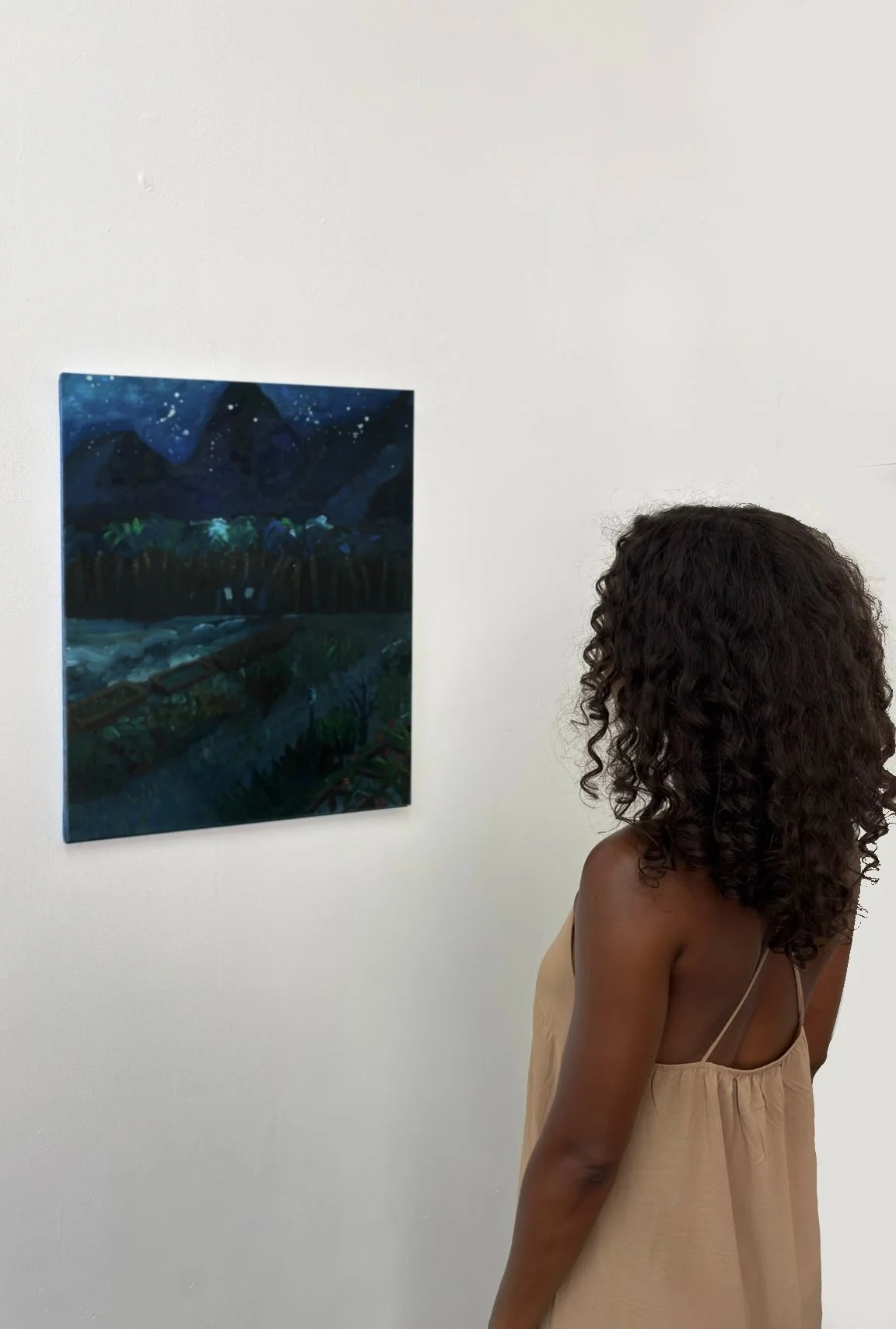 A woman with curly hair in a beige dress looking at a dark landscape painting in a gallery.