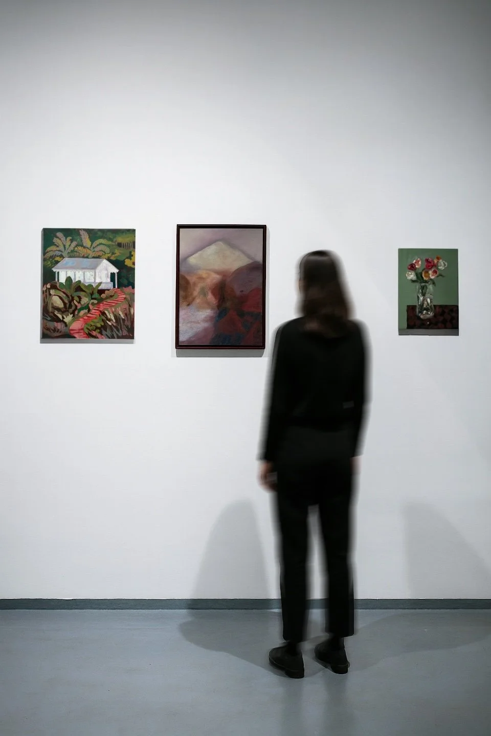 A woman with dark hair wearing a black outfit stands in an art gallery, observing three paintings on a white wall.