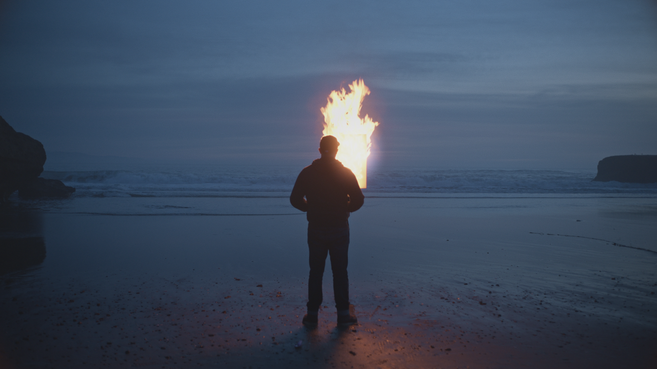 A person standing on a beach at night, facing a large burning structure or object near the ocean.