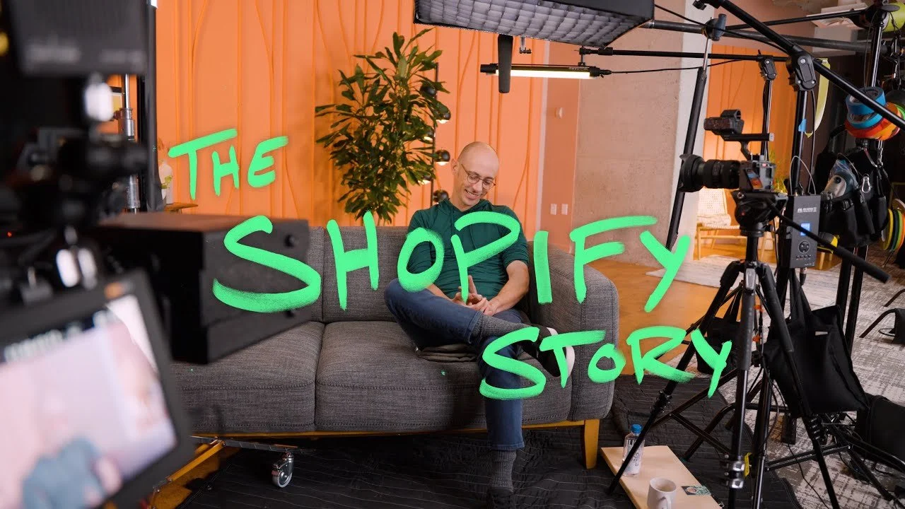 A man sitting on a gray couch with a green background and the words 'The Shoify Story' written on it, surrounded by camera equipment in a recording studio.