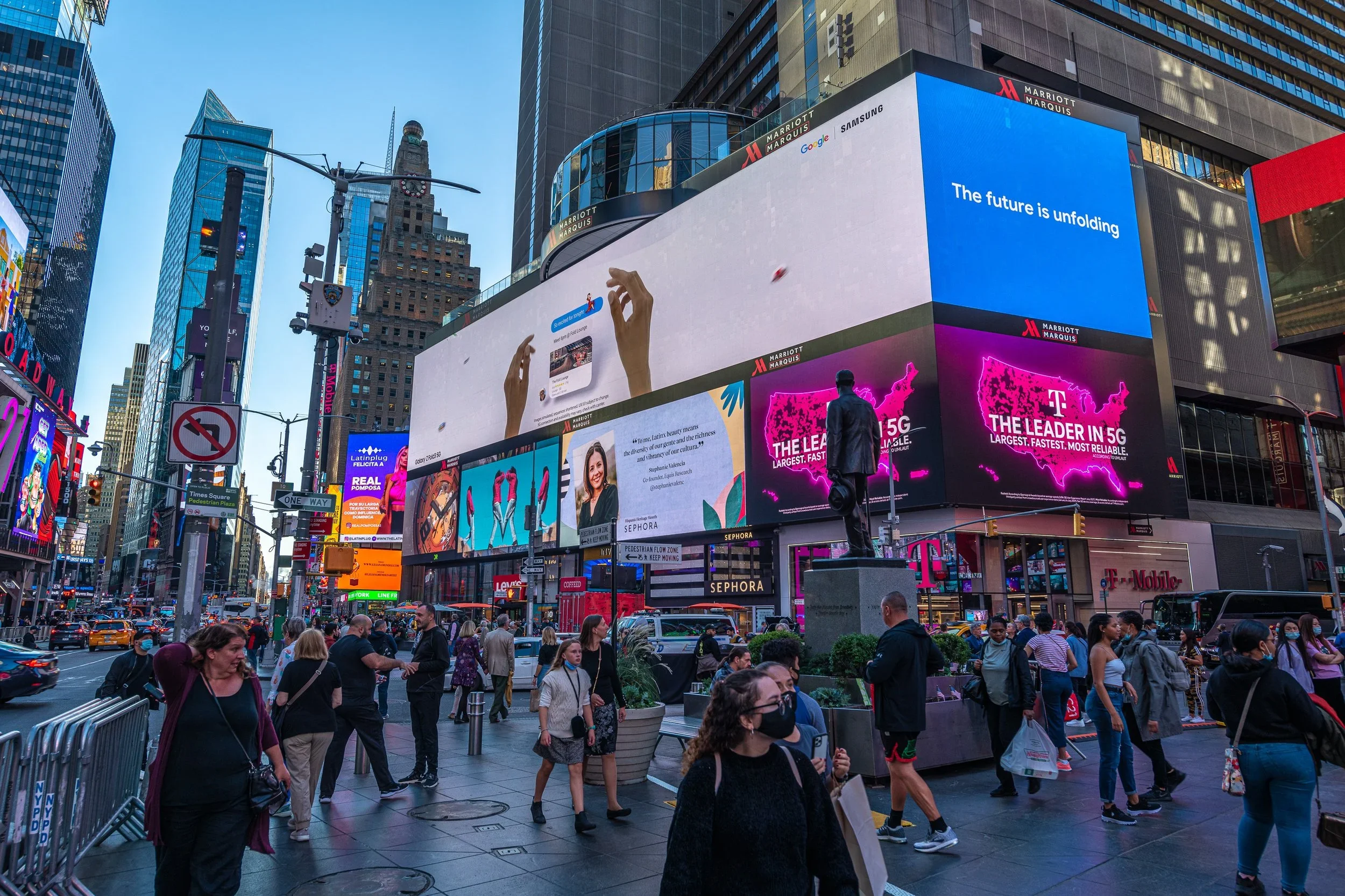 Crowded Times Square in New York City with large digital billboards, pedestrians crossing the street, and yellow taxis in traffic.