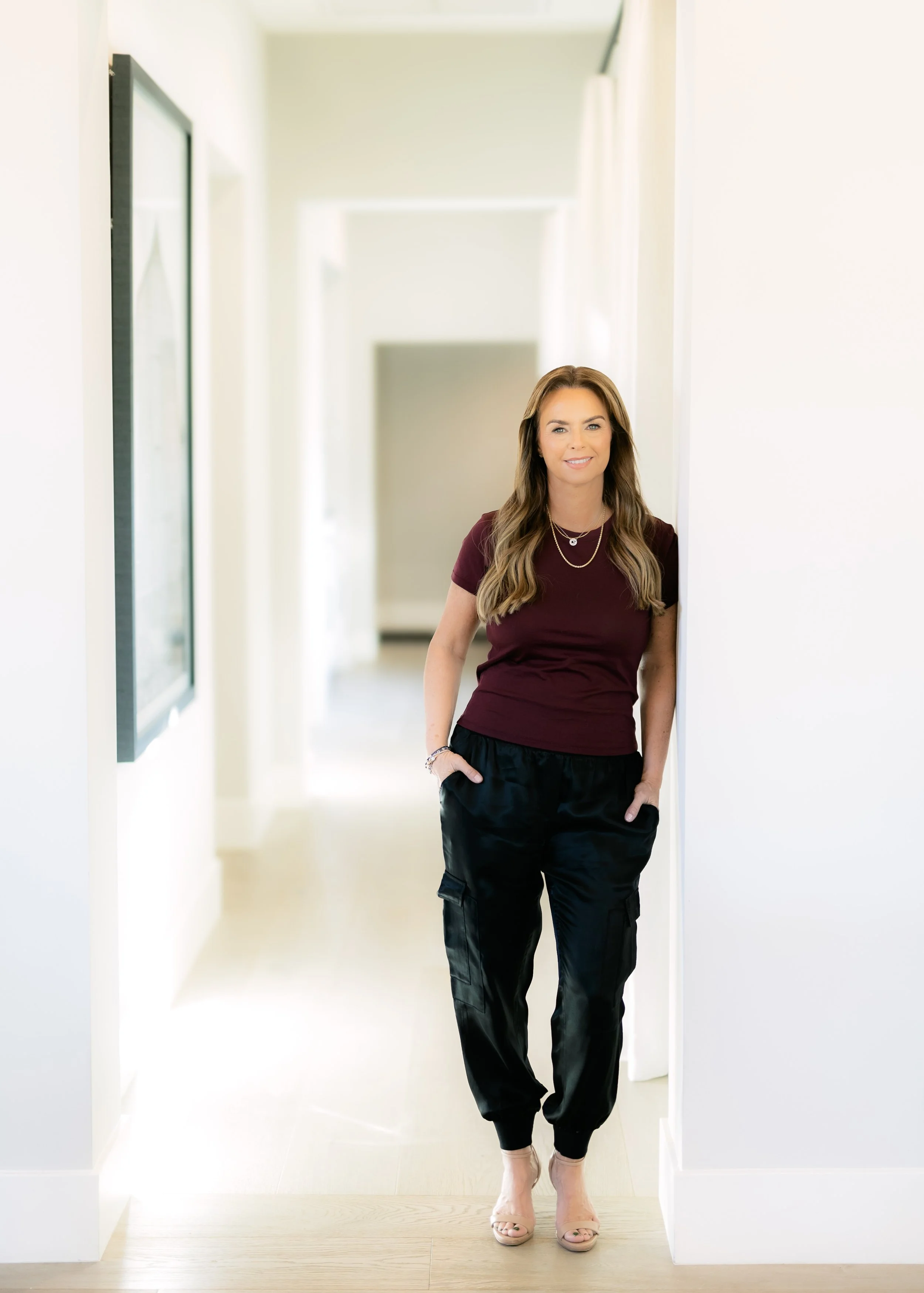Char Sarraf standing in a brightly lit hallway, wearing a maroon t-shirt, black cargo pants, and beige heels, smiling at the camera.