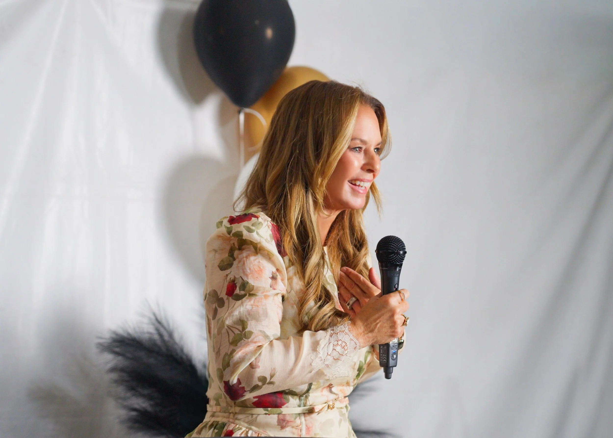 Keynote Speaker, Char Sarraf, with long blonde hair holding a microphone, smiling, and wearing a floral dress, standing in front of white backdrop with black, gold, and white balloons.