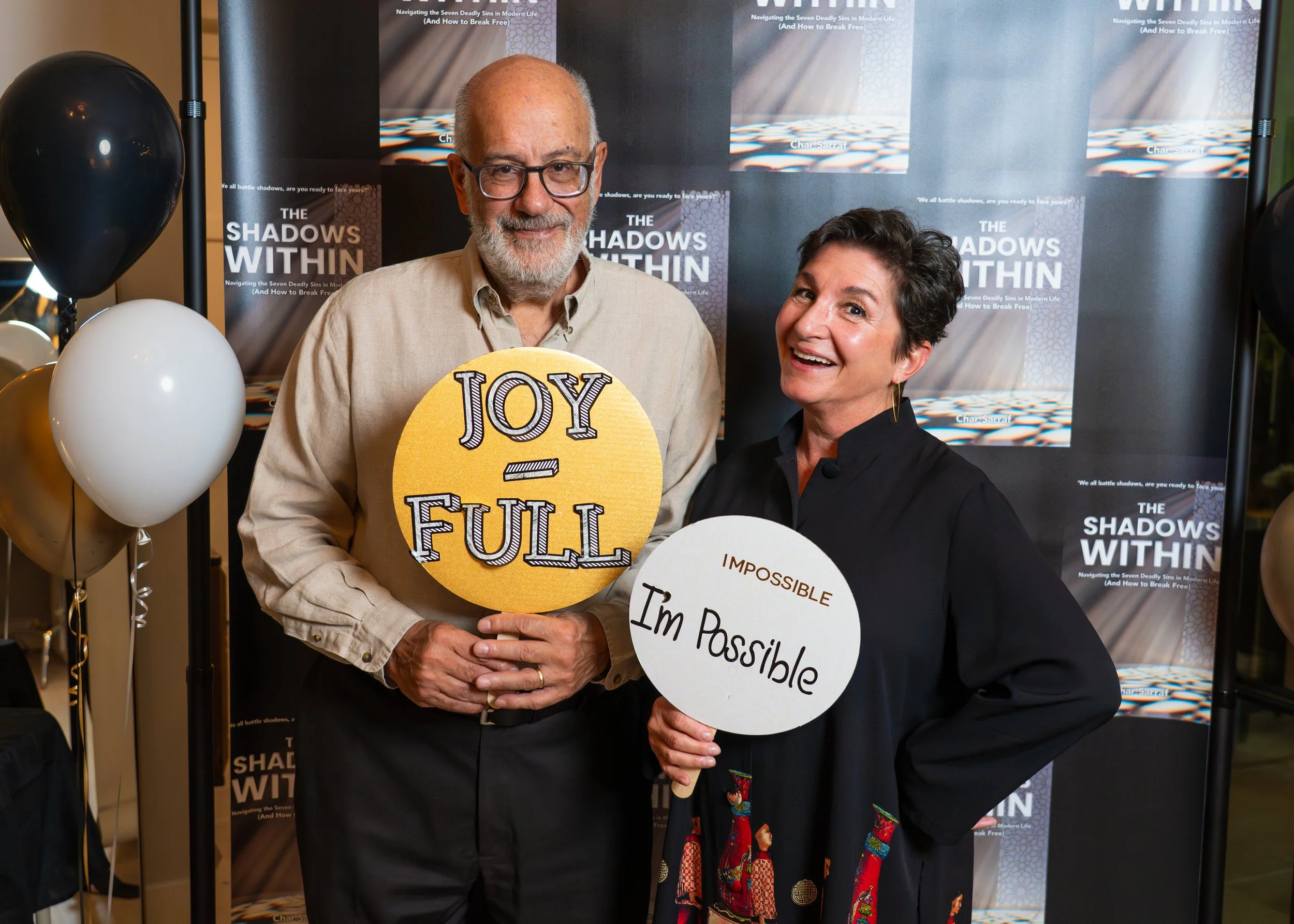 A man and a woman smiling and holding signs, standing in front of a backdrop with Char Sarraf's book covers. The man's sign says 'Joy Full,' and the woman's sign says 'Impossible I'm Possible.'