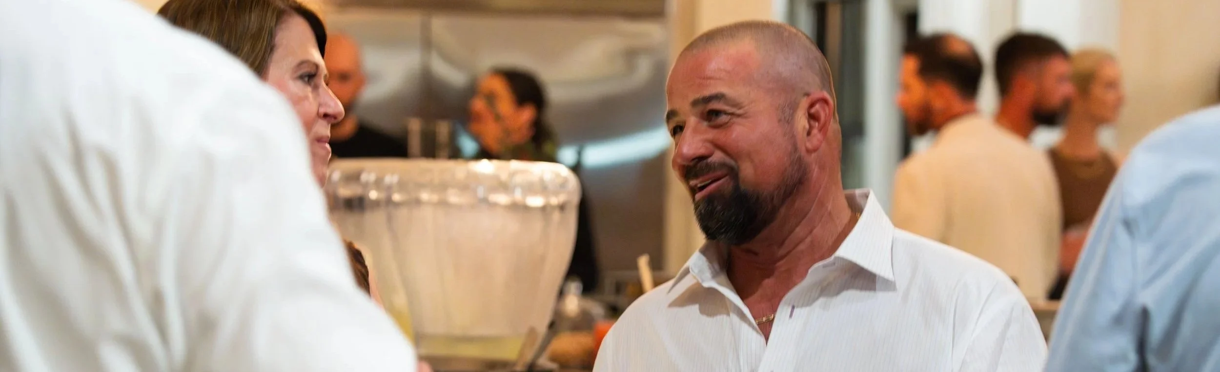 A man with a beard and bald head wearing a white shirt smiling and talking to a woman at Char Sarraf's Speaking Event with other people in the background.