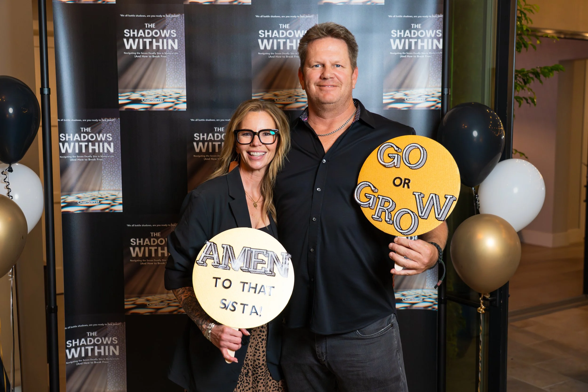 A man and woman smiling at Char Sarraf's speaking event. The woman has blonde hair, glasses, and tattoos on her arm, and the man has short hair and is holding a sign that reads "GO OR GROW".
