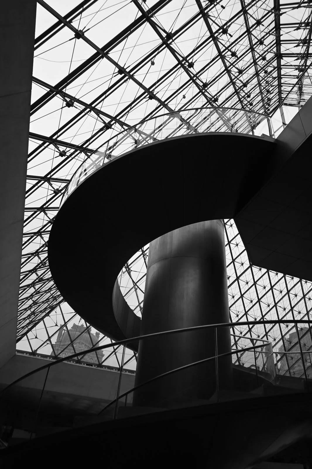 Low-angle black and white photograph of the spiral staircase beneath the glass pyramid at the Louvre Museum in Paris, France.