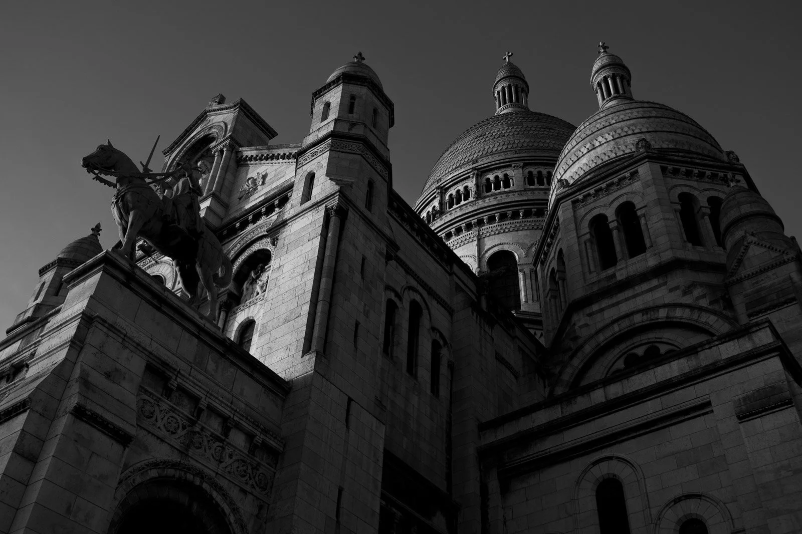 Low-angle black and white photograph of the equestrian statue of Joan of Arc with the Sacré-Cœur Basilica rising prominently behind her in Montmartre, Paris, France.
