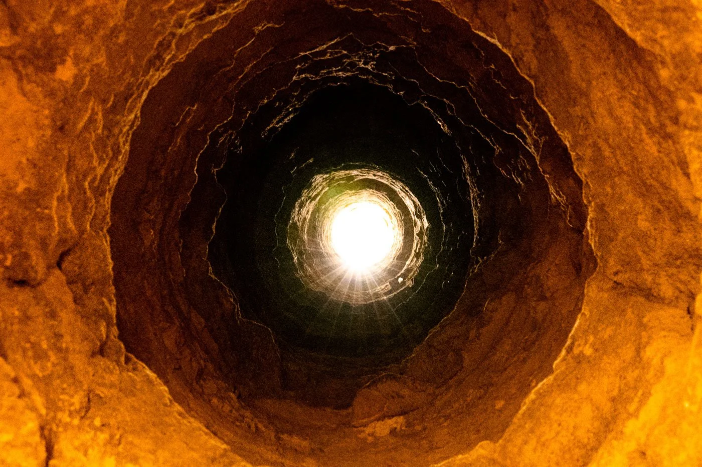 Upward view from the bottom of a vertical tunnel in the Paris Catacombs showing warm orange stone walls, dark silhouetted midsection, and bright sunlight at the opening above in Paris, France.