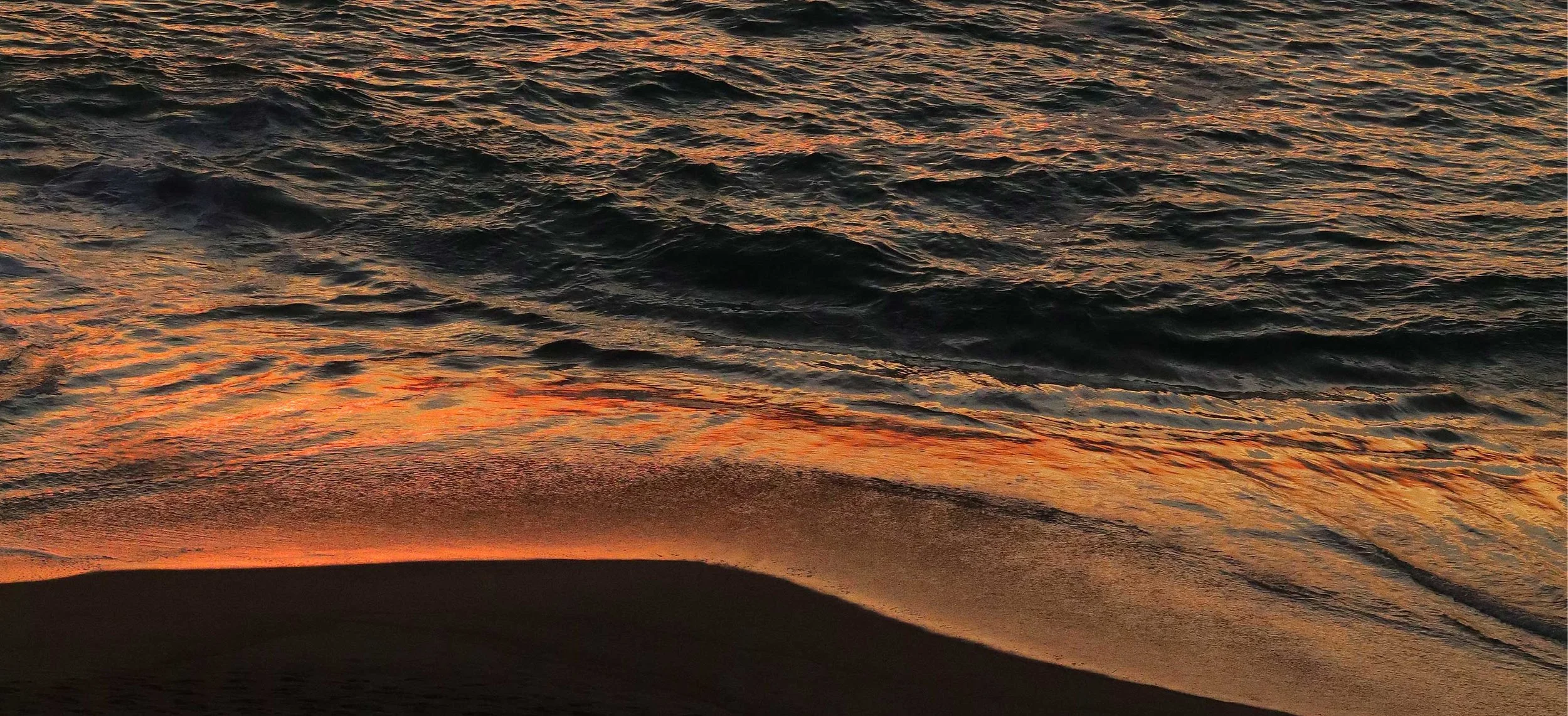 The photo shows a sunset over the ocean, with waves reflecting orange and gold hues from the sky, and a part of the sandy beach in the foreground.