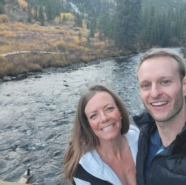 A smiling couple taking a selfie near a river with a forested hillside in the background.